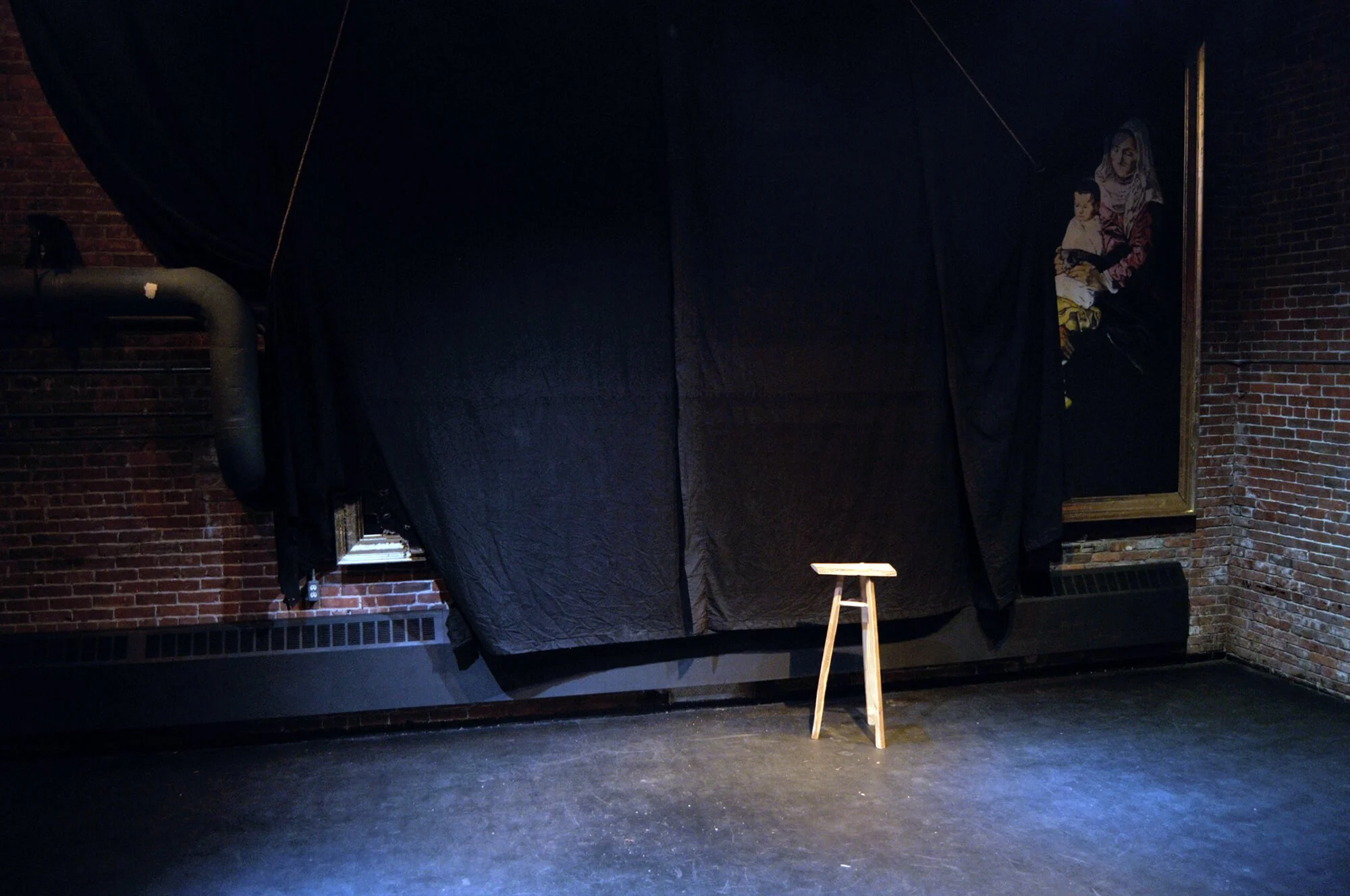 Empty stool on stage with black curtain, brick wall, and a painting of a woman with children in the background.