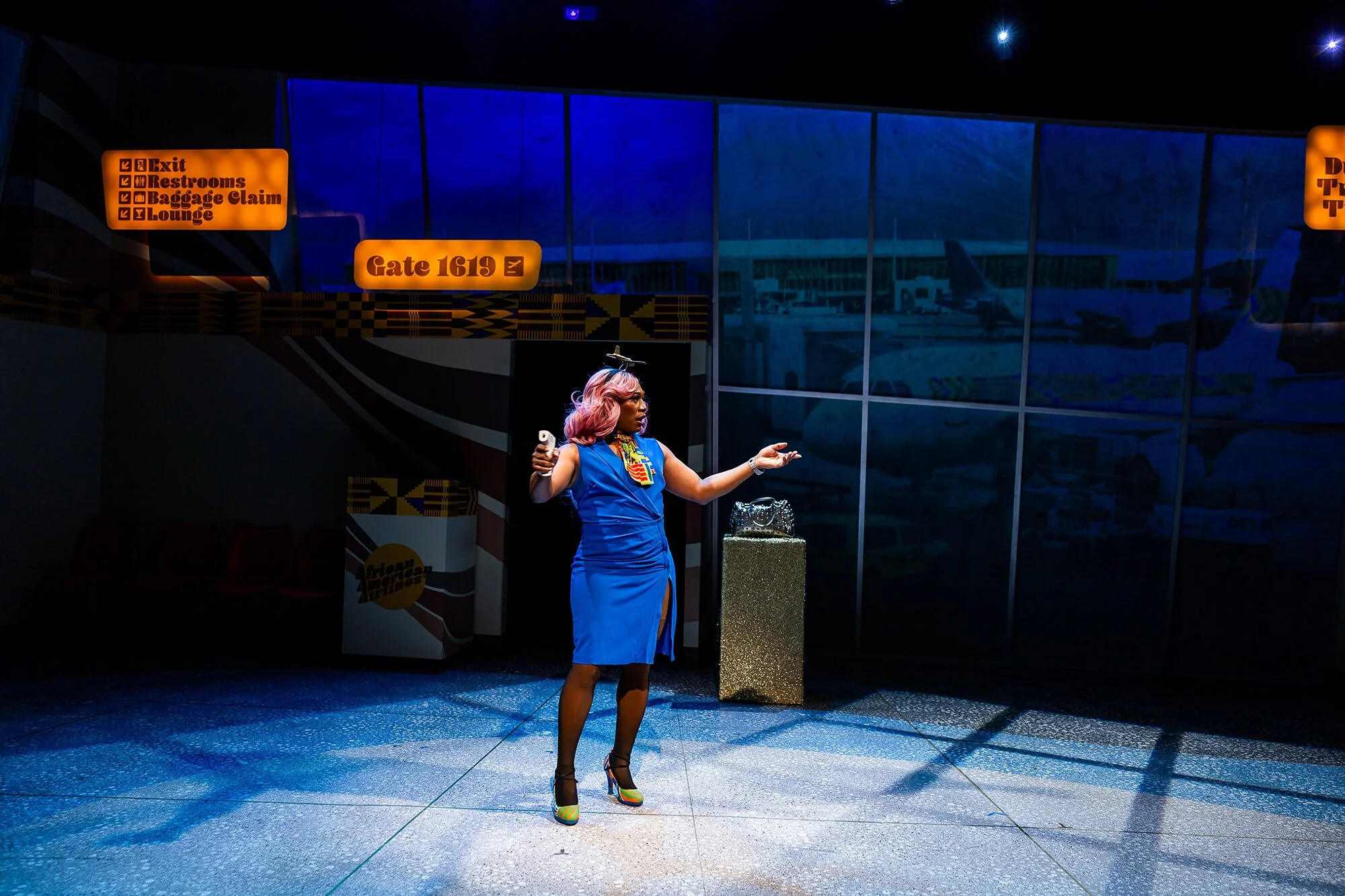 A woman with pink hair wearing a blue dress and colorful high heels is standing in an airport terminal, holding a phone in one hand and gesturing with the other. There are orange signs overhead with directions and a glass wall reflecting airplanes ou