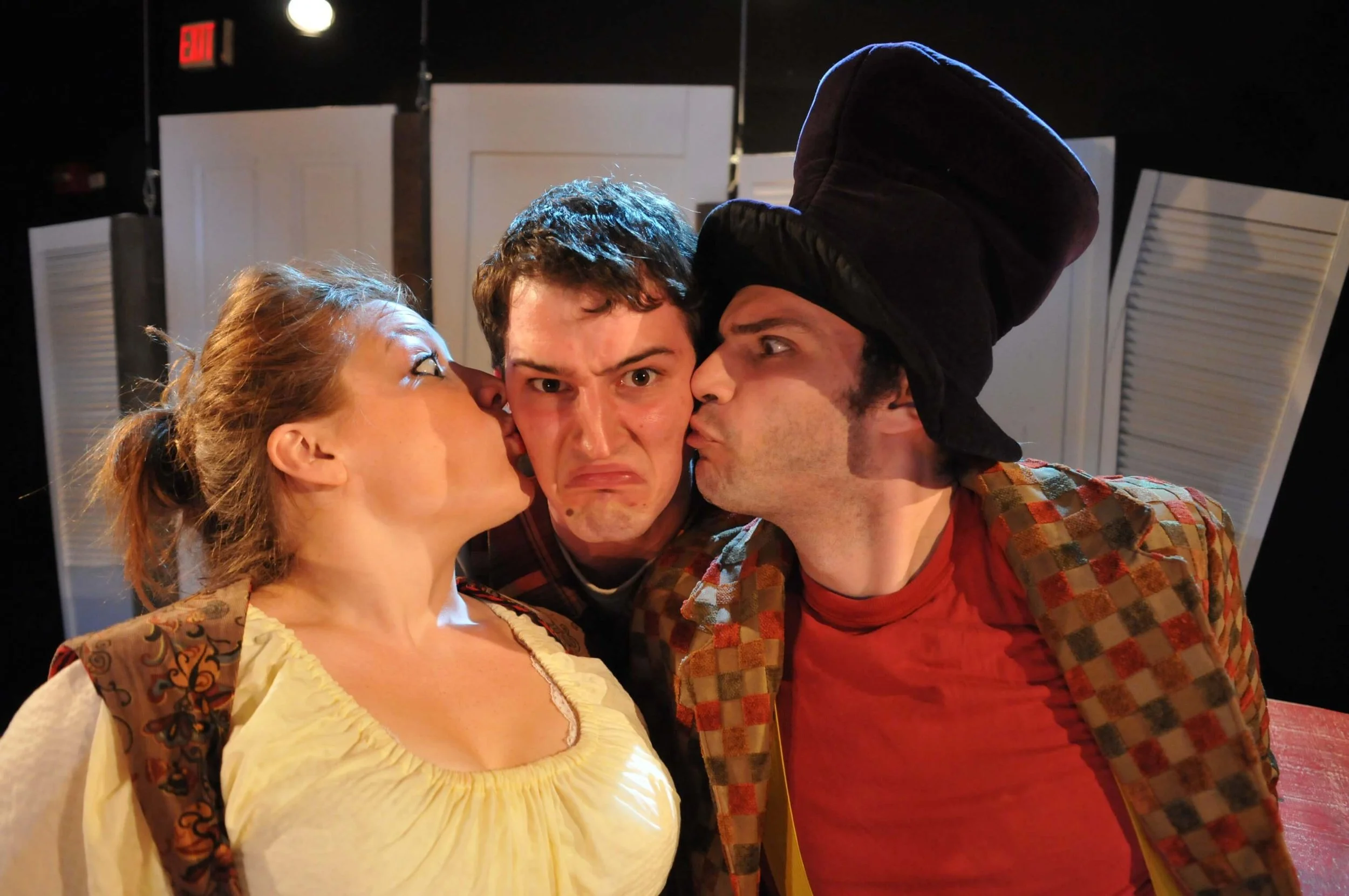 Three people on stage, two men and one woman, with one man in the middle looking uncomfortable while the other two kiss him on the cheeks.