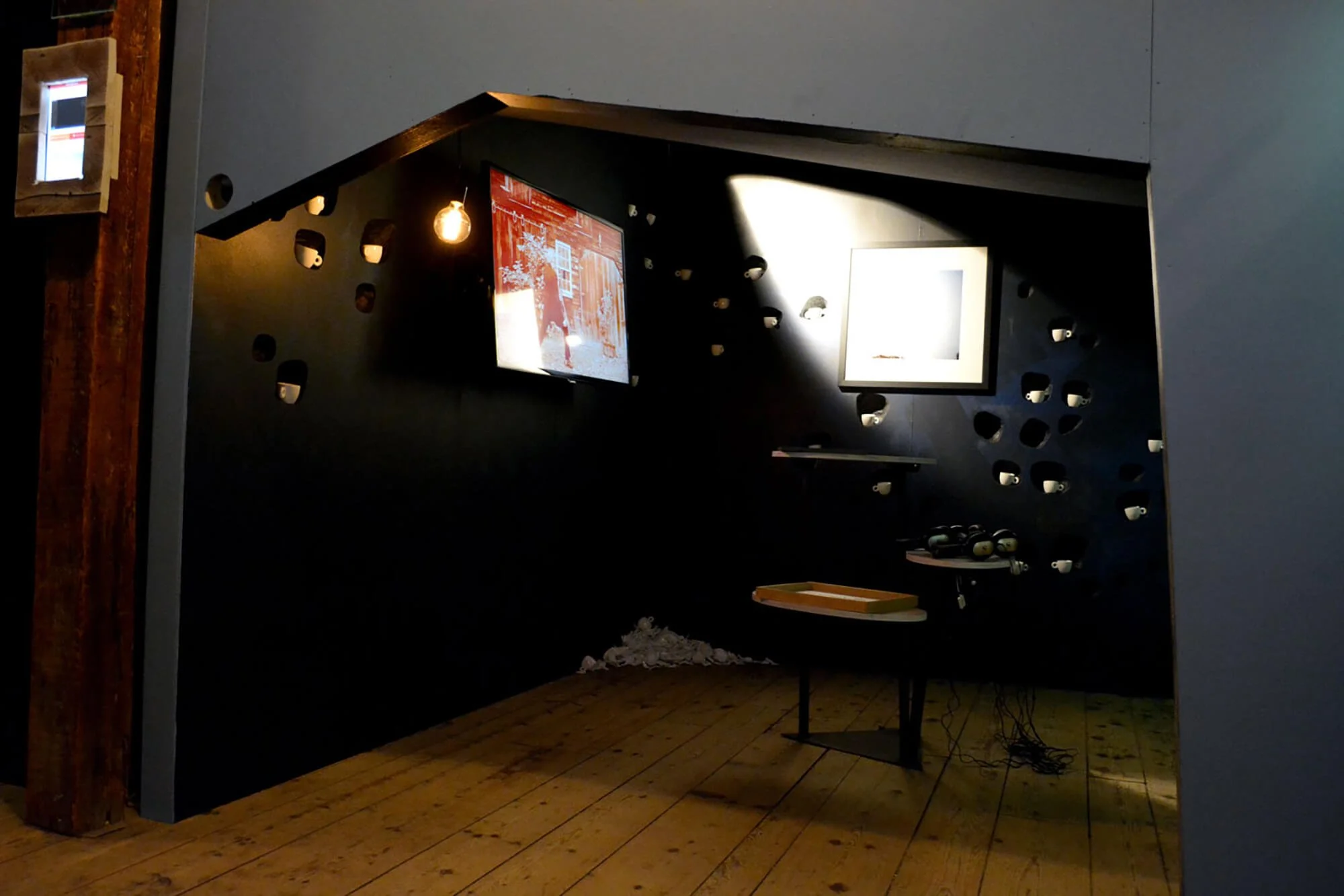 An art installation featuring two screens on a dark wall, with cups embedded around them, wooden flooring, and a small table with objects on it, illuminated by a single exposed light bulb.