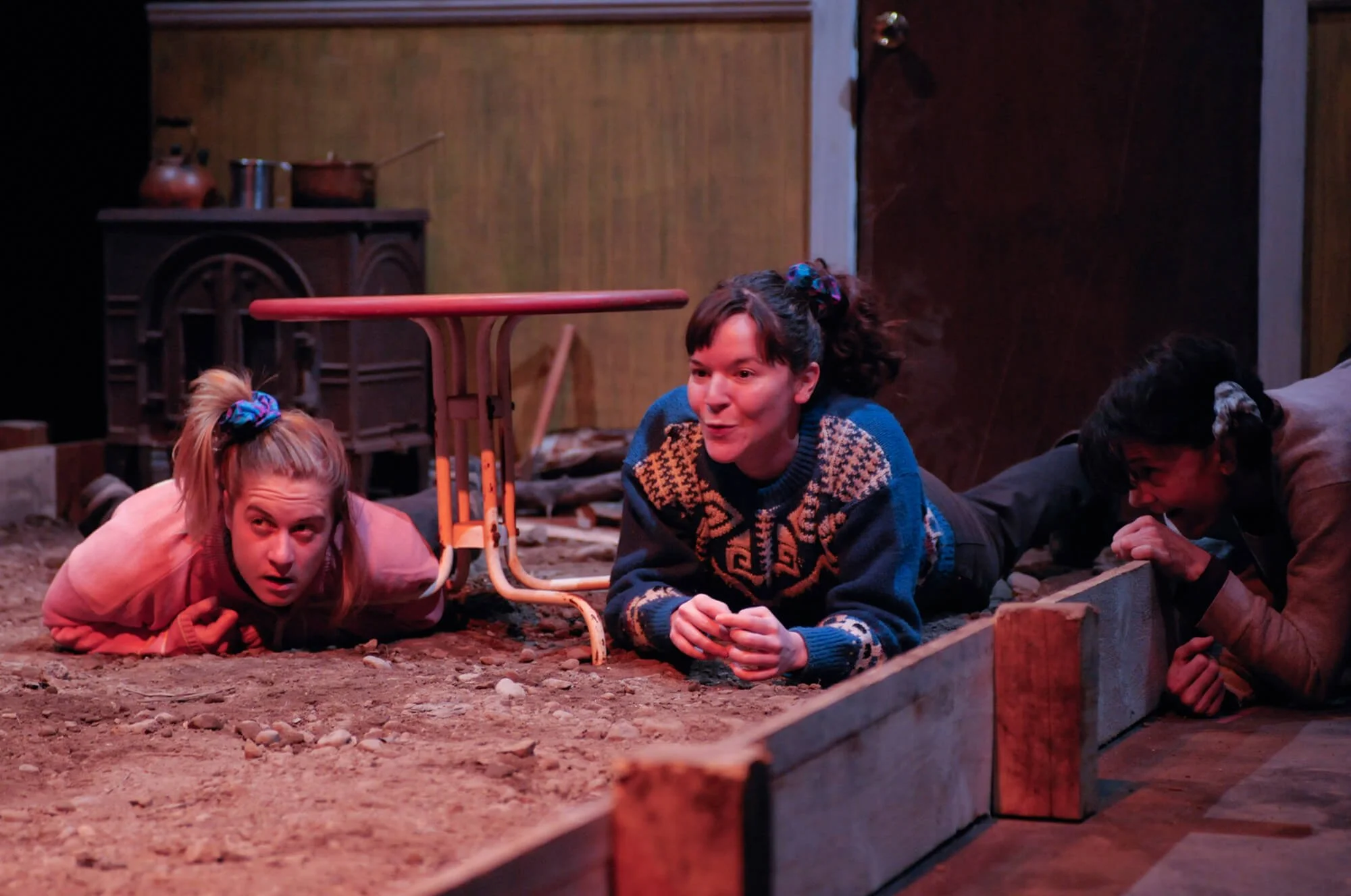 Three women lying on the ground under a table and against a wooden wall, engaged in a theatre scene. One woman on the left with blonde hair in a ponytail, wearing a pink jacket, looks upward with a surprised expression. The woman in the middle with d
