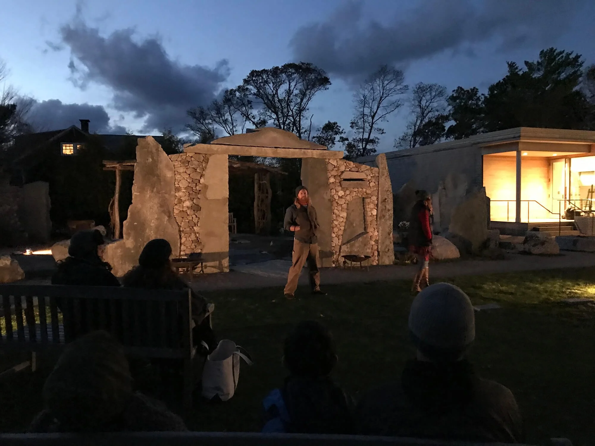 People sitting on benches and standing outdoors at dusk, watching a small performance near stone and wood structures, with trees and a darkening sky in the background.