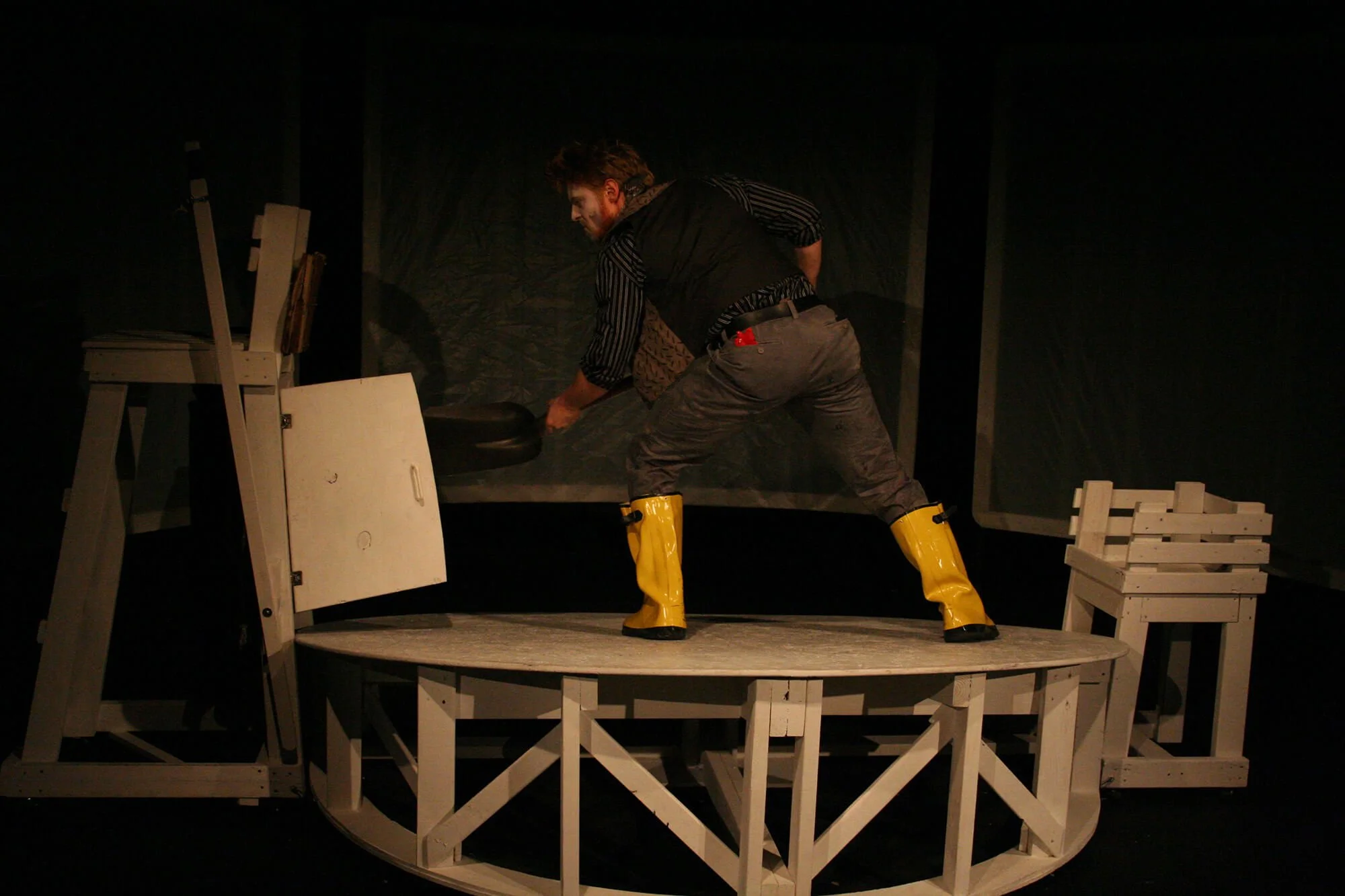 A man with a beard in theatrical costume, wearing yellow boots, is lifting a black circular object on a stage with minimal scenery, including a white chair and a small white bench, in a dark setting.