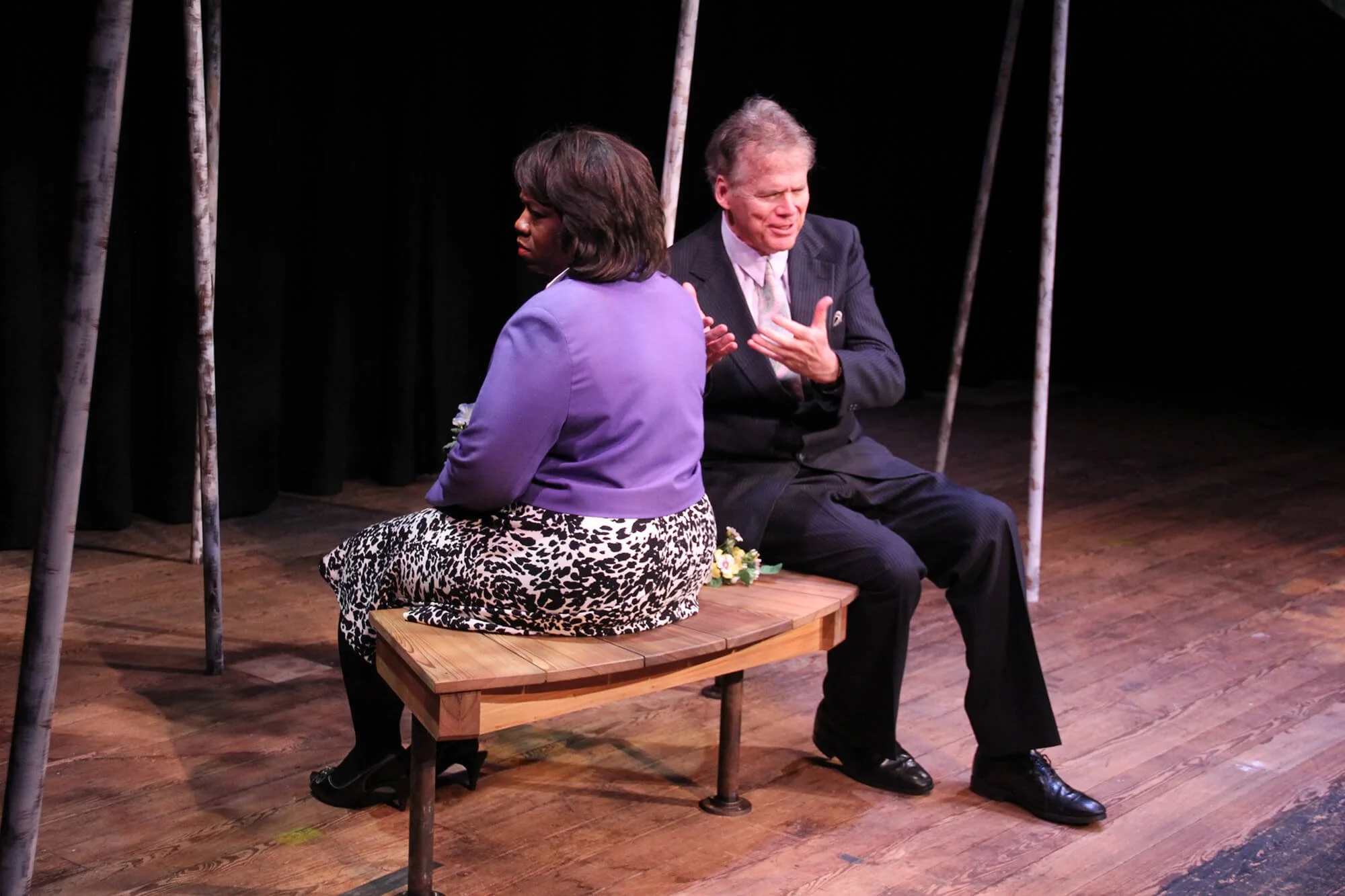 Two actors sitting on a wooden bench on a stage, with black curtains and standing poles in the background, engaged in a theatrical performance.