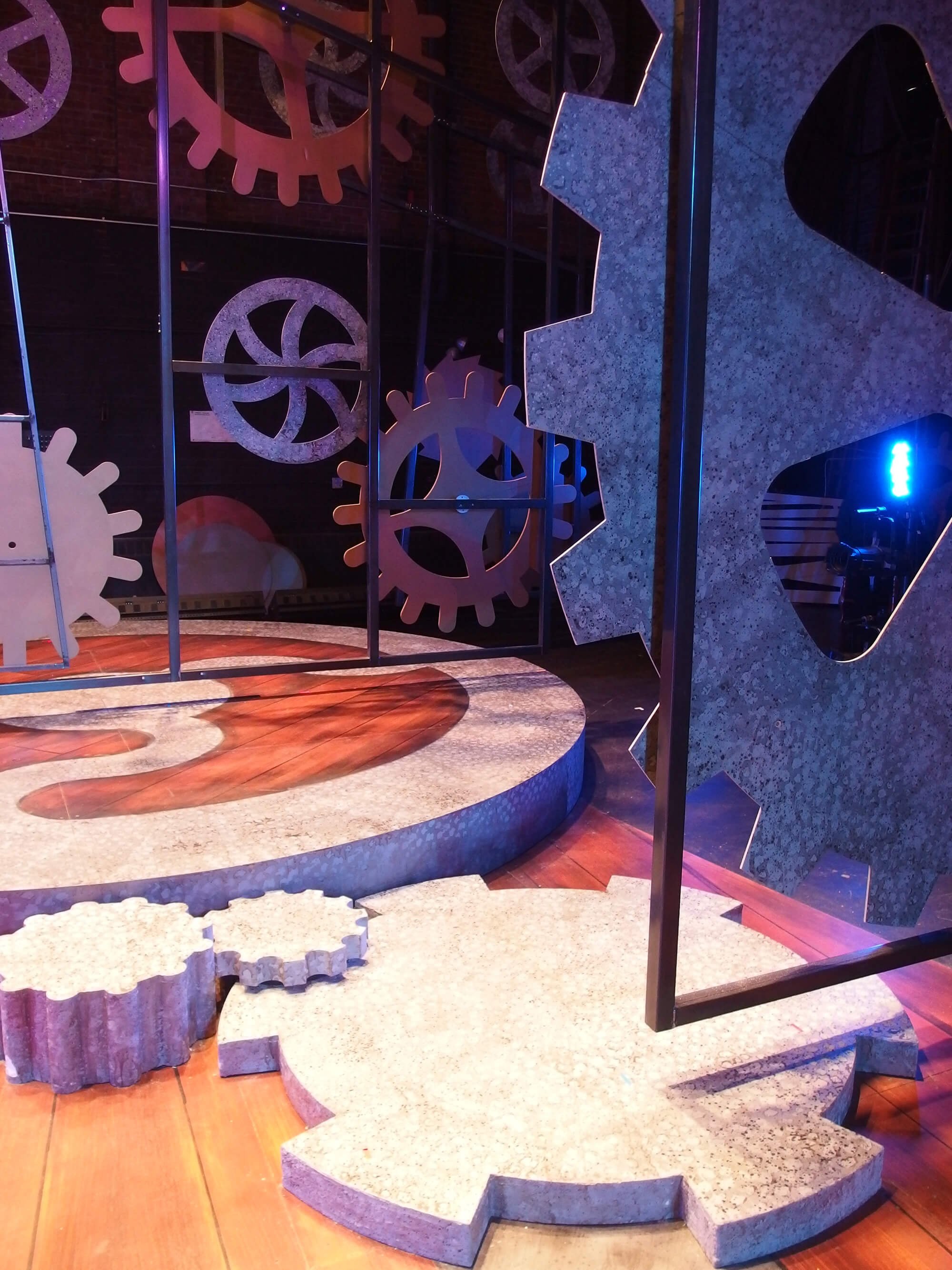 The image shows a mechanical-themed exhibit with large gears and cogs made of stone and metal, set on a wooden floor with a colorful, industrial-style background.
