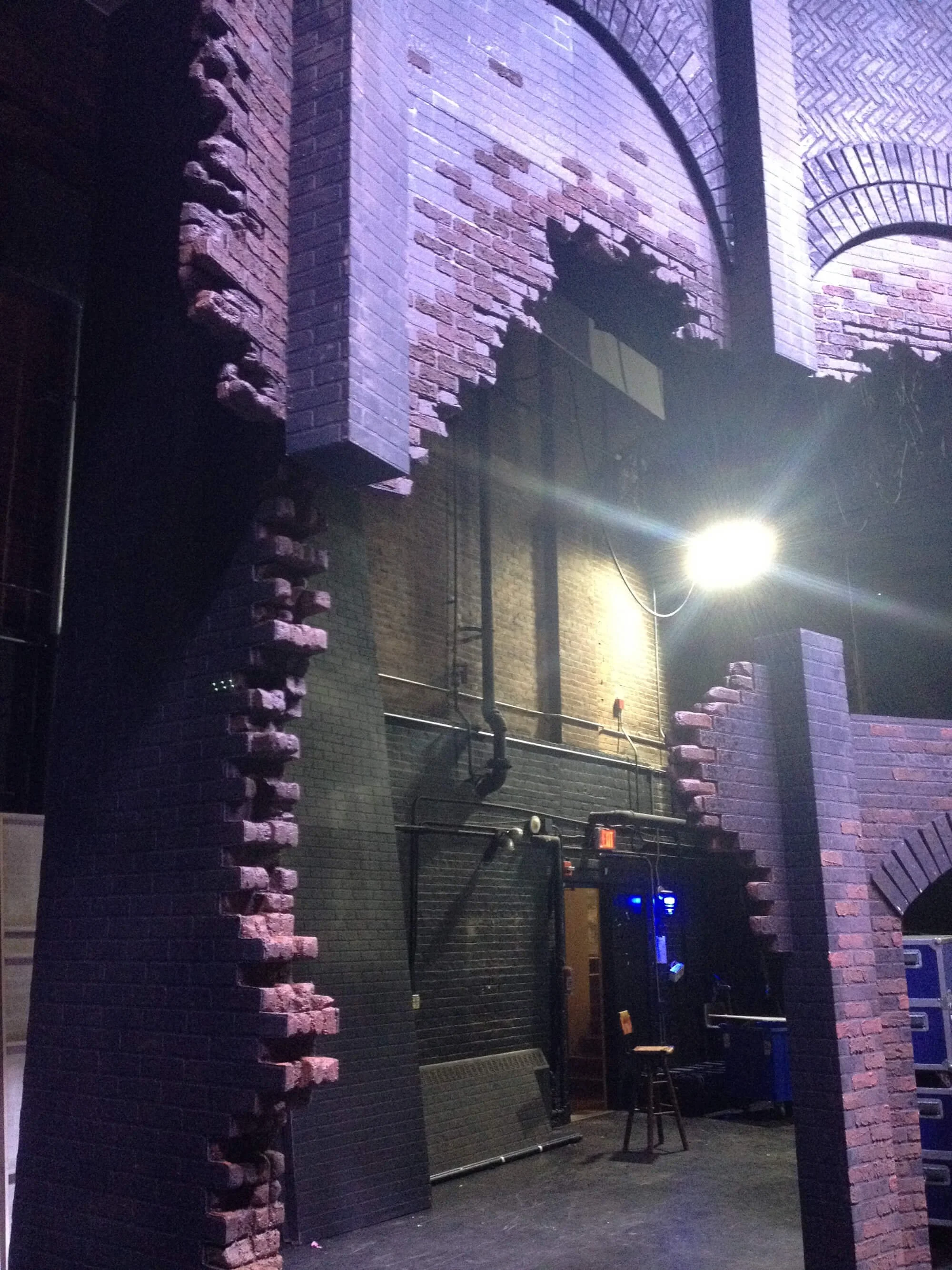 Nighttime view of a brick building with large arched windows, illuminated by a bright outdoor light. The wall features exposed pipes and a doorway with a stool next to it.