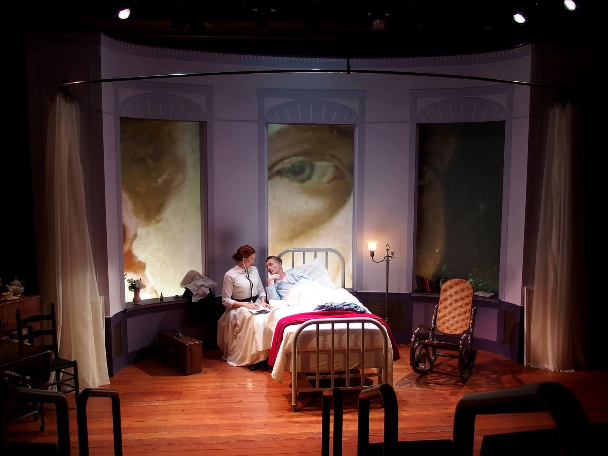Stage set resembling a bedroom with large projected eyes on the back wall. Two actors, a woman and a man, are sitting on a bed engaged in conversation. The woman is dressed in period costume, and the man is lying down, looking up at her. Surrounding 