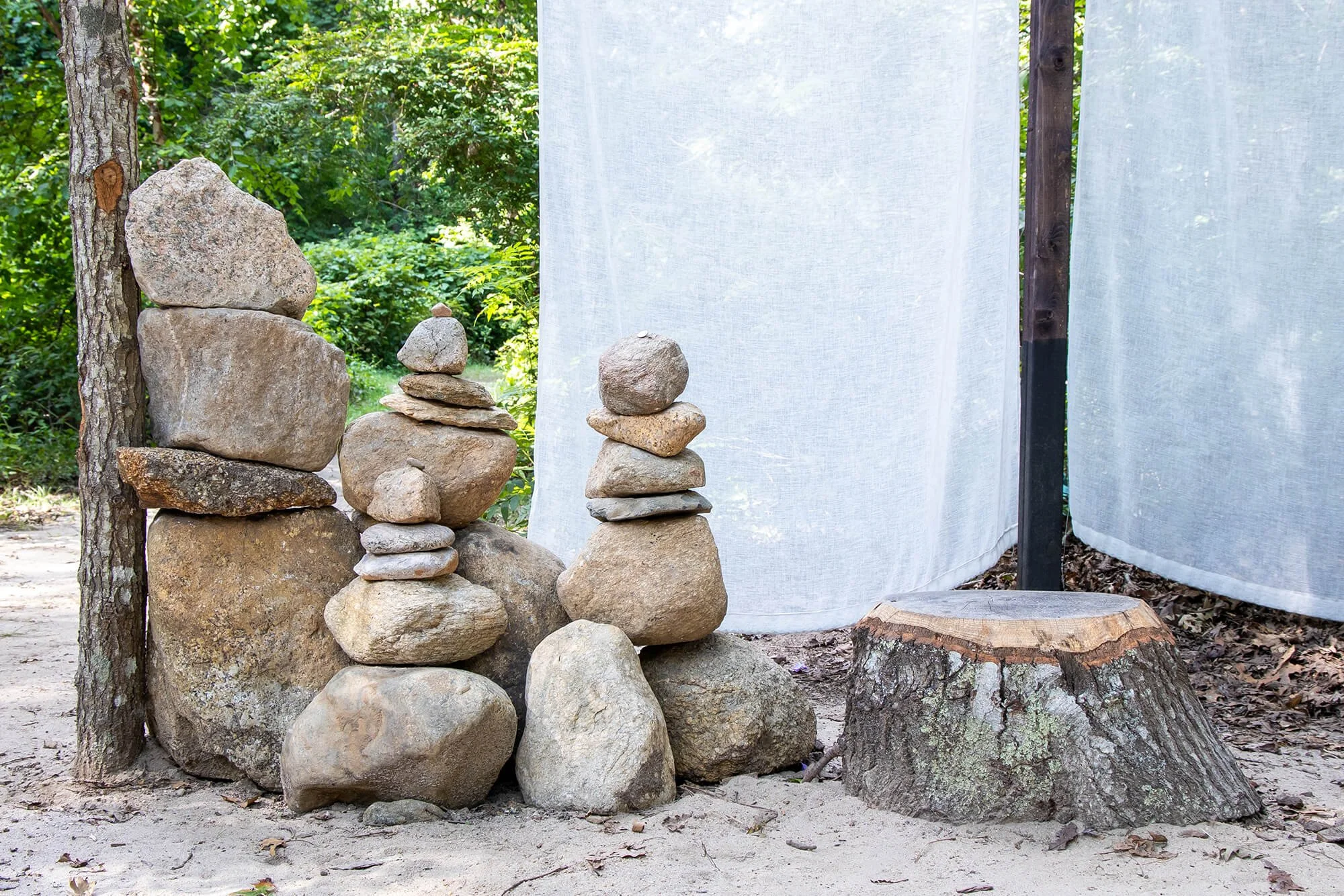 Stacks of rocks arranged on the ground near a tree and white fabric backdrop in an outdoor wooded area.