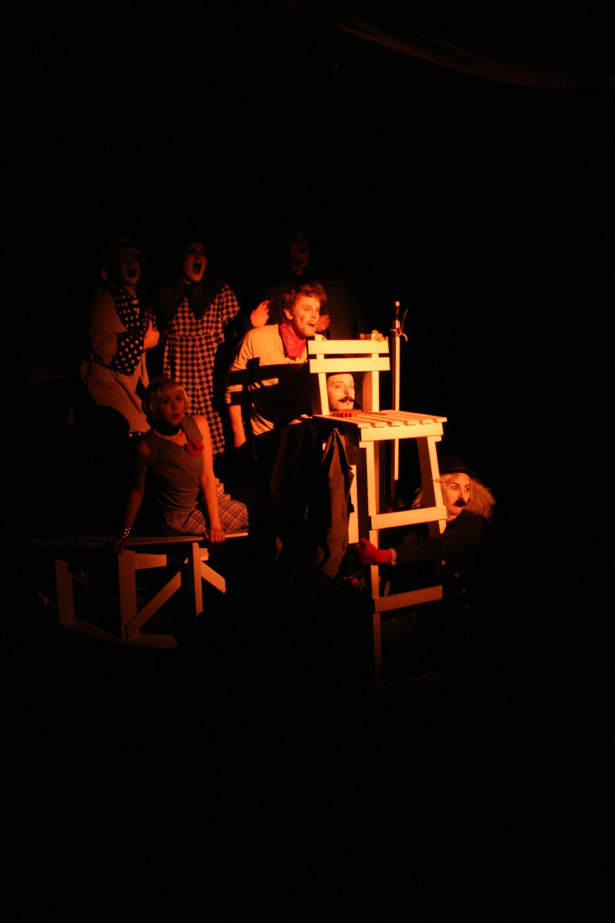 A theatrical stage performance with actors in clown makeup and costumes, some on a wooden structure, in a dark setting