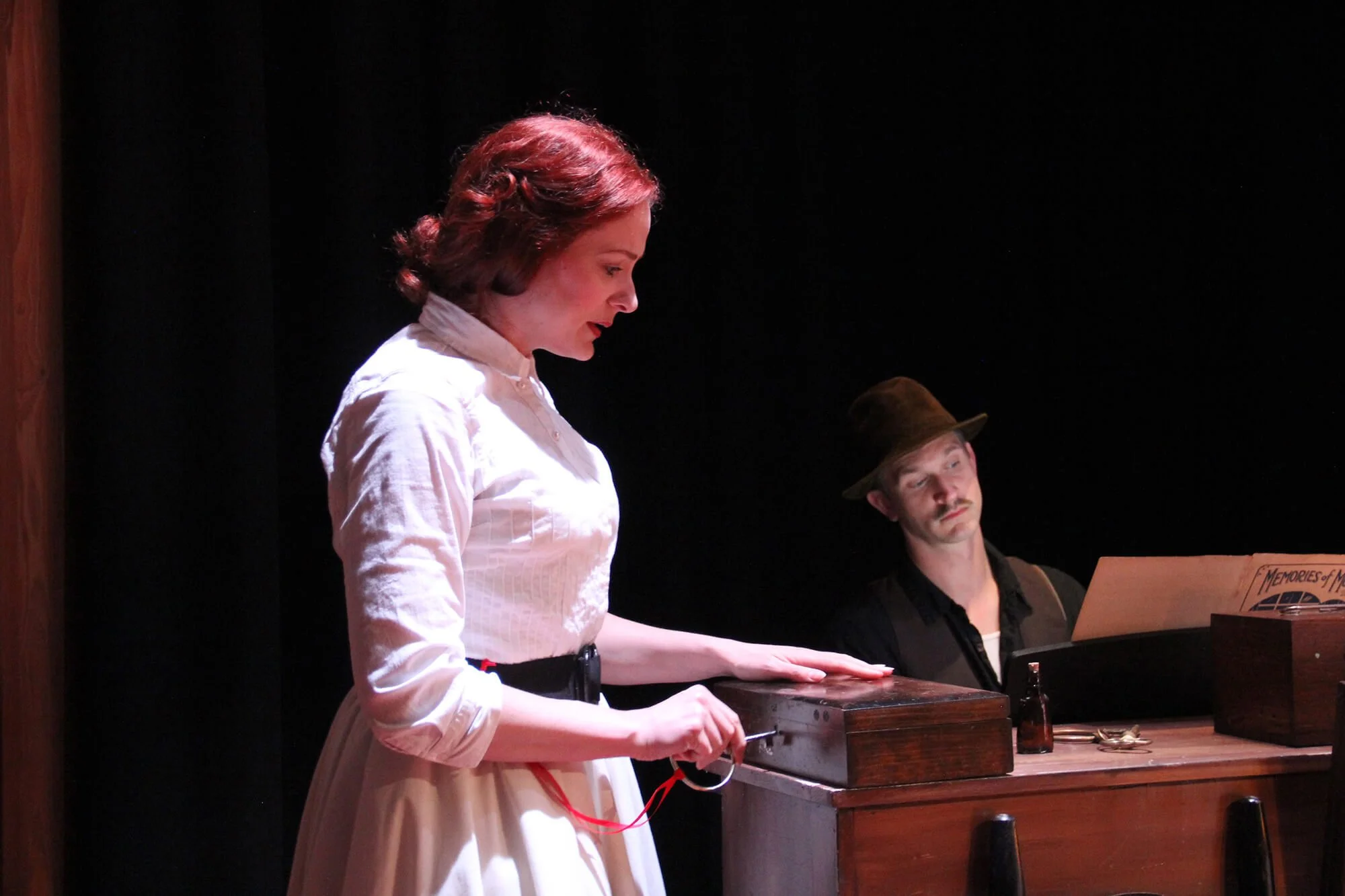 A woman with red hair, dressed in a white blouse and skirt, appears to be in a theatrical performance, looking down with a contemplative expression. A man wearing a brown hat and dark clothing sits beside her, looking at her with a serious expression