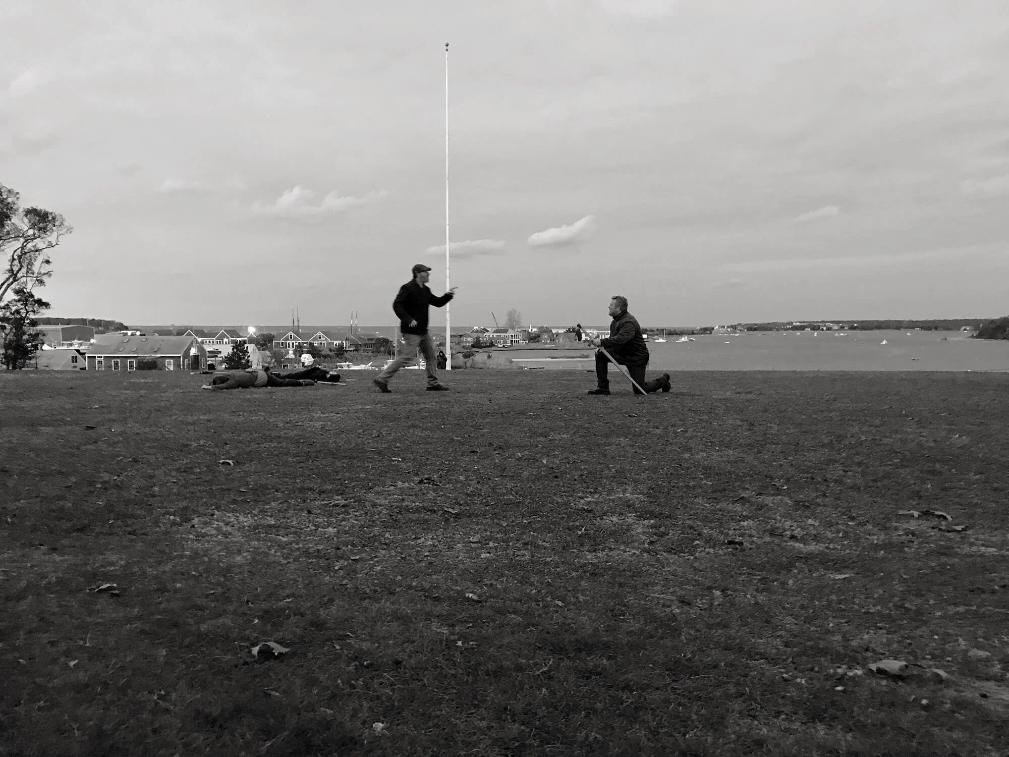 Two men are on a grassy area near water, one standing with a long flagpole, the other kneeling holding a stick. Two people are lying down in the background, and a small town or neighborhood is visible across the water.