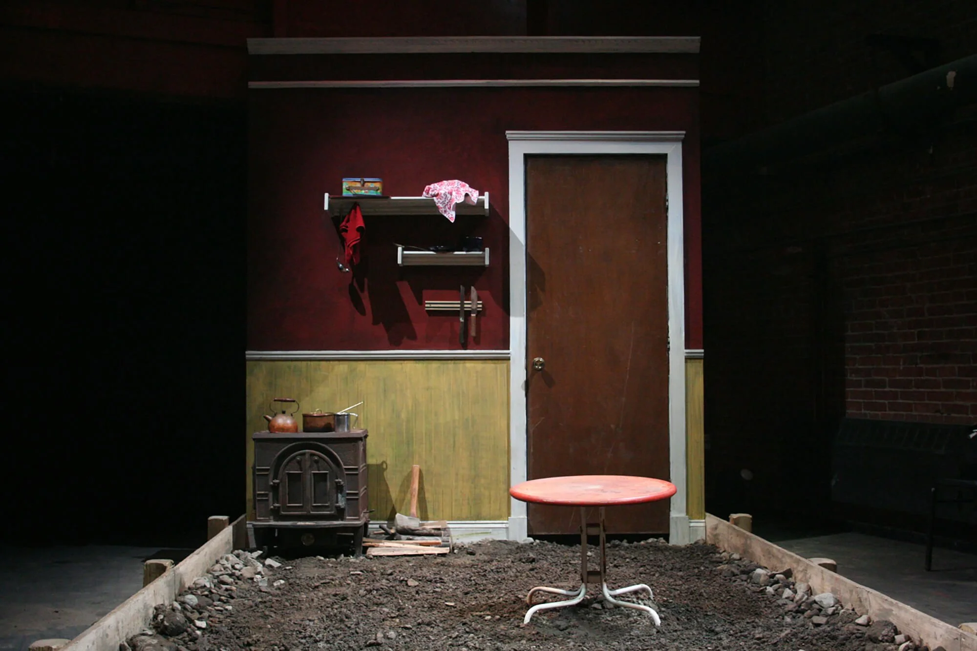 A small theatrical set depicting a simple room with a closed wooden door, a mounted shelf with clothing and miscellaneous items, a stove with pots, and a small round table with a red top on a dirt-covered floor.