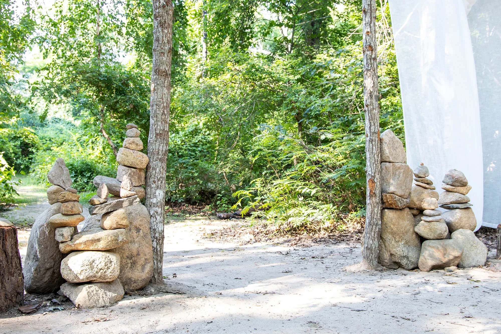 Two small stone stacks, or cairns, are placed between two trees in a wooded outdoor area. One on the left, and one on the right of the image. In the background, lush green trees and foliage are visible, and the ground appears sandy or dusty.