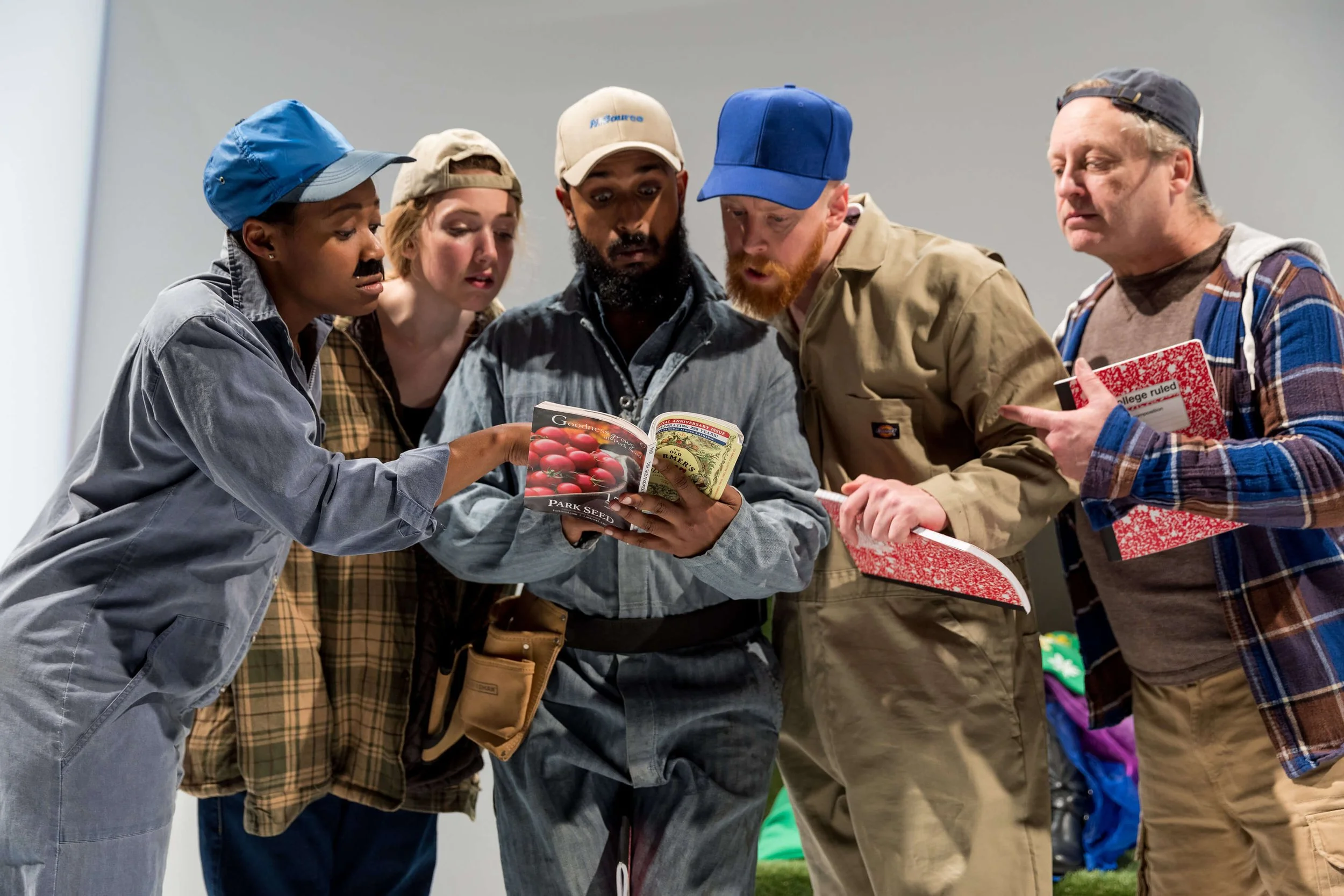 Group of five diverse people gathered closely, looking at a book about gardening or plants, with some holding notebooks or tablets, in an indoor setting.
