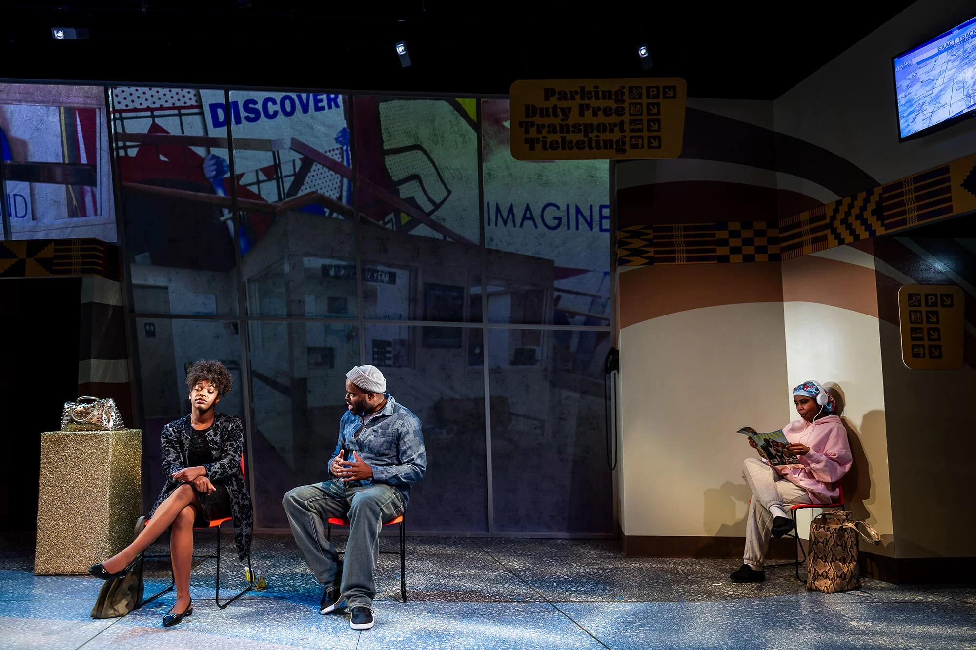 Two people sitting and talking in a waiting area, with a third person reading a magazine nearby, in an indoor setting with a large wall backdrop and signs for parking, duty-free, transport, and ticketing.