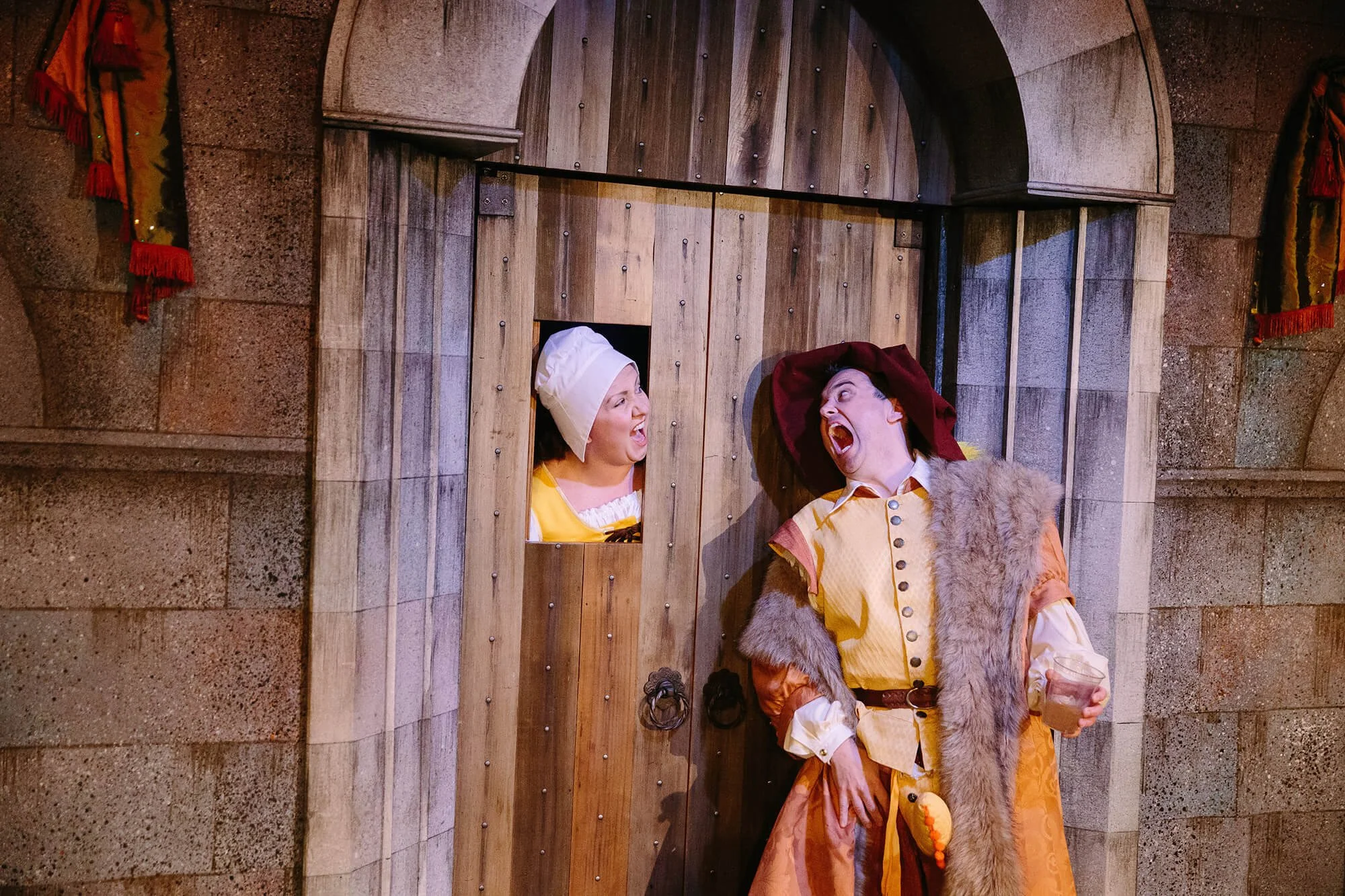 Two actors in costume, one woman with a chef's hat and another man with a red turban, engaging in an animated conversation through a small window in a wooden door surrounded by stone walls, suggesting a theatrical performance.