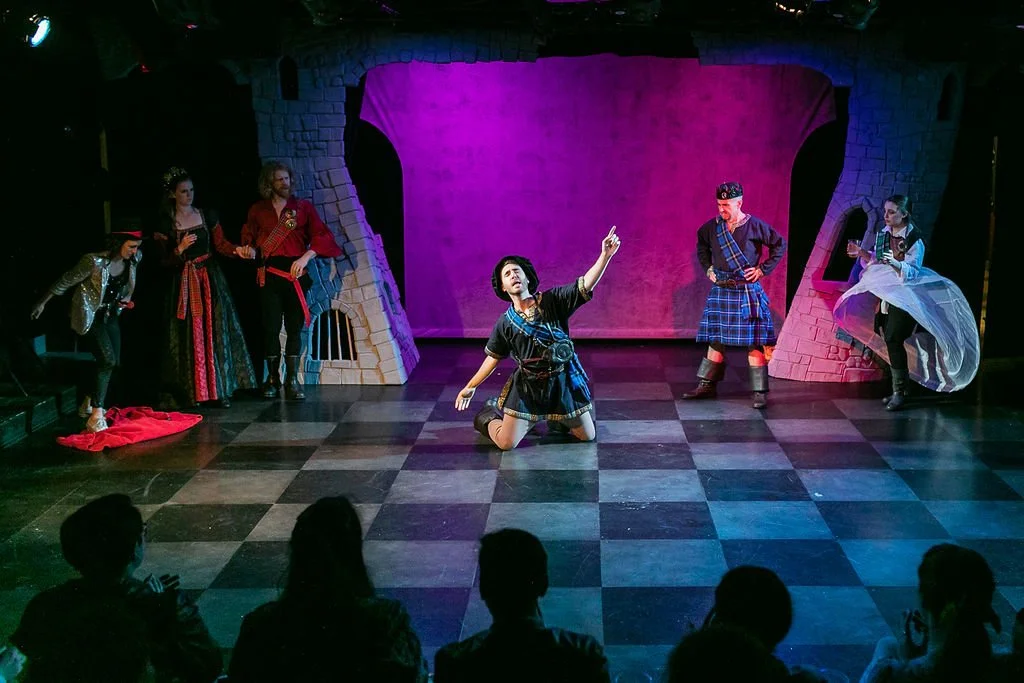 Actors performing a theatrical play on stage with a checkered floor, set with a pink backdrop and stone archways, with a small audience watching.