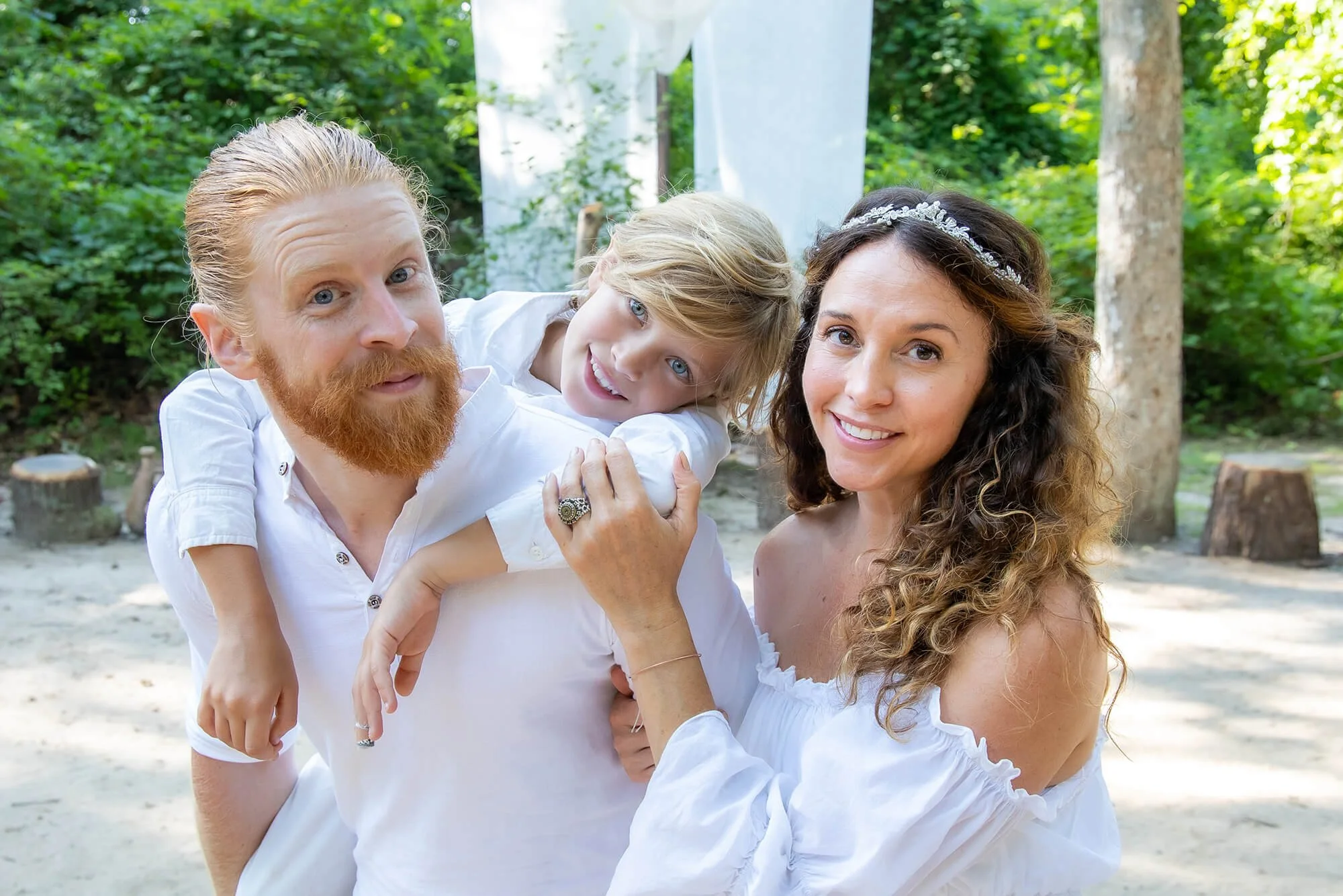 A family of three, a man with red hair and beard, a woman with curly hair, and a young boy with blond hair, smiling outdoors surrounded by trees and greenery.