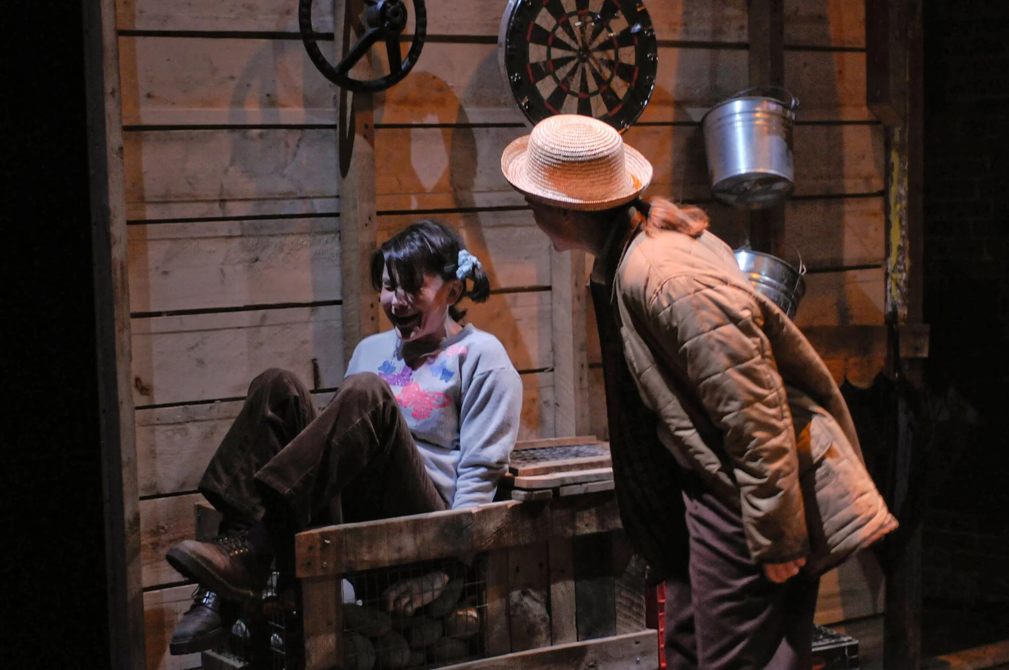 A girl sitting laughing inside a wooden shack with a woman leaning toward her. The girl has dark hair with a blue bow, wearing a gray sweatshirt and brown pants. The woman is wearing a straw hat, brown quilted jacket, and has reddish hair. A dartboar