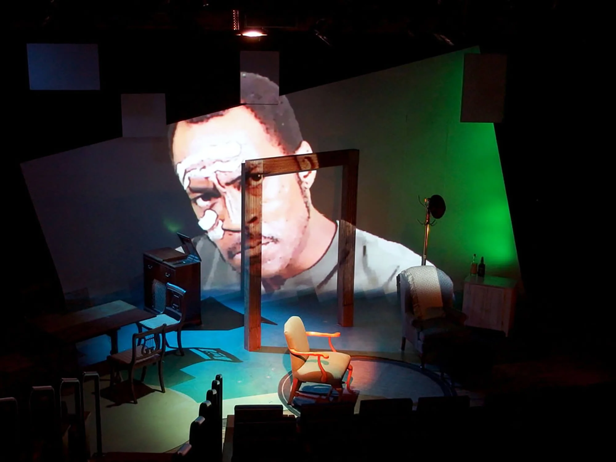 Stage set with a large projection of a man's face, surrounded by chairs and furniture, with colorful lighting.