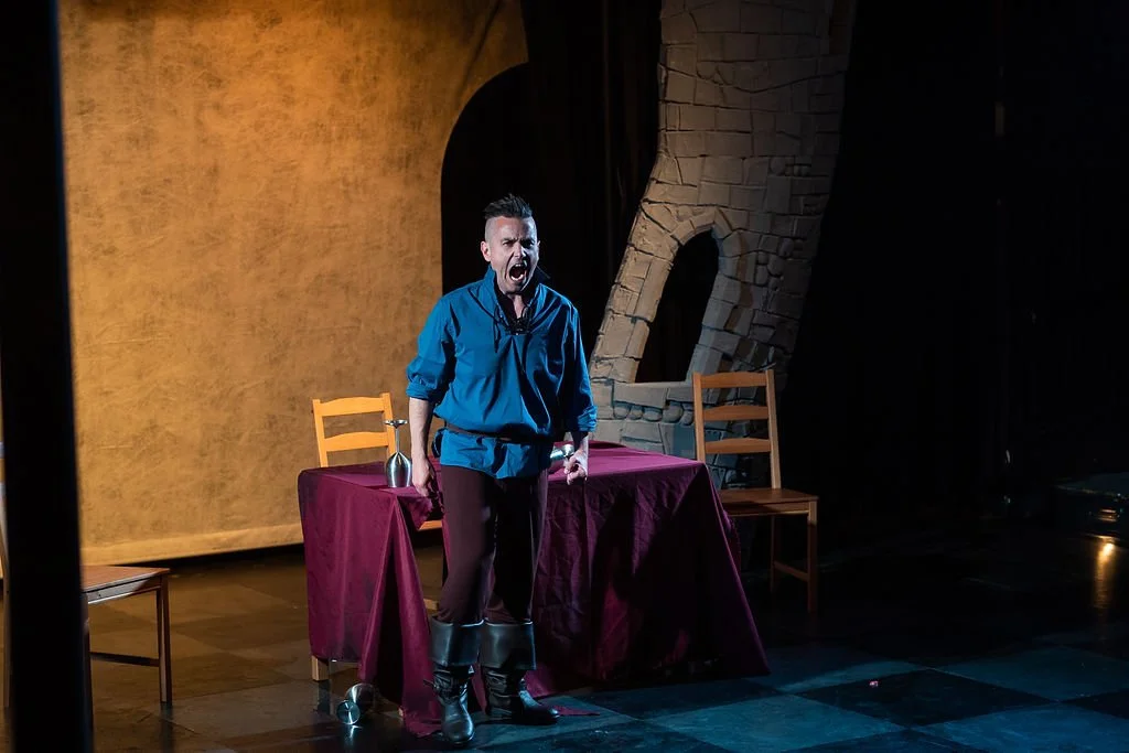 A man is on stage, yelling or singing passionately, wearing a blue shirt, dark pants, and black boots. There is a table covered with a maroon cloth behind him, with a wine bottle and a glass on it. Two wooden chairs are beside the table. The backgrou
