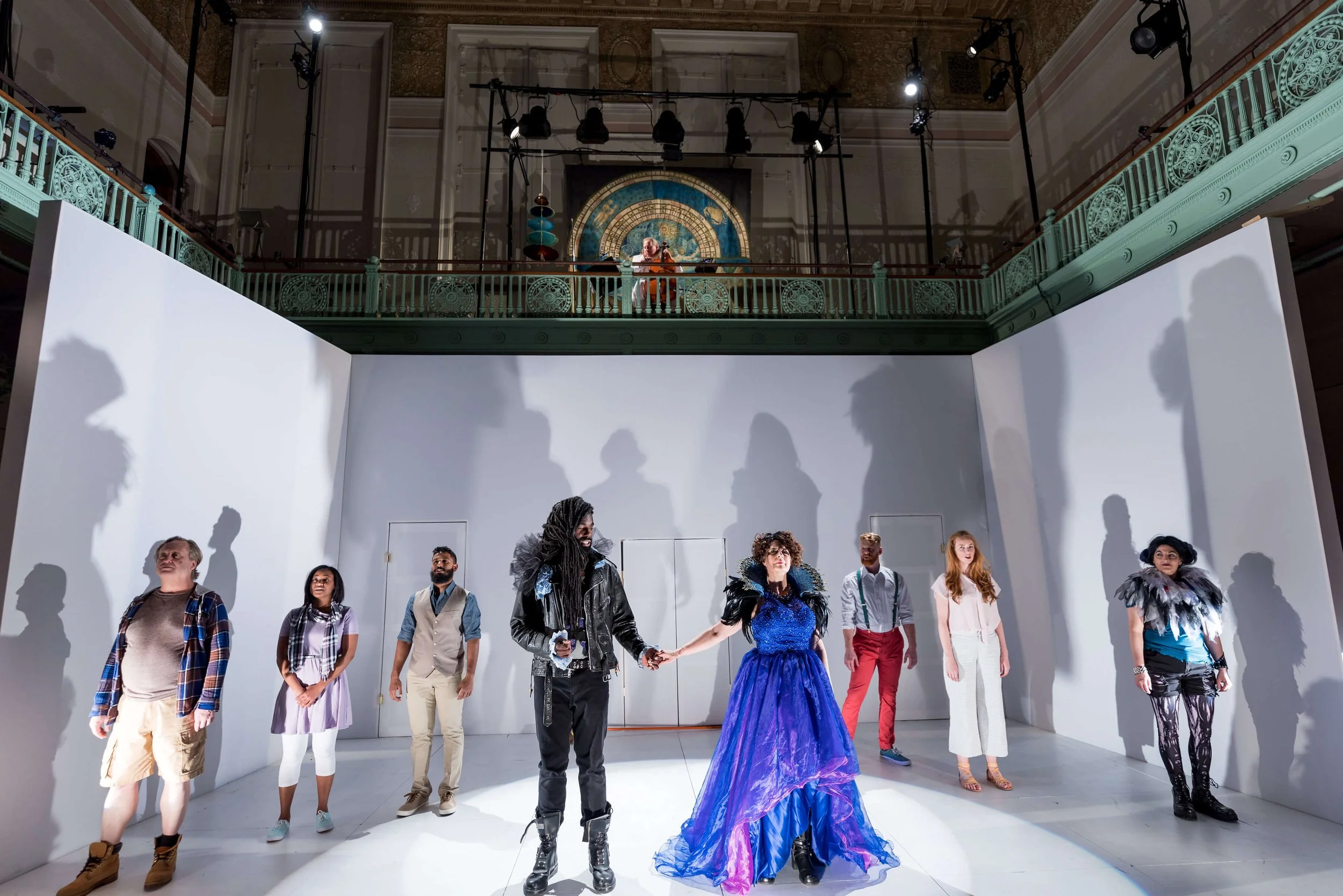 A theatrical stage scene with nine actors standing in a line on white stage with white walls, gesturing toward each other, with a backdrop of shadows, and a balcony above with two more actors observing.