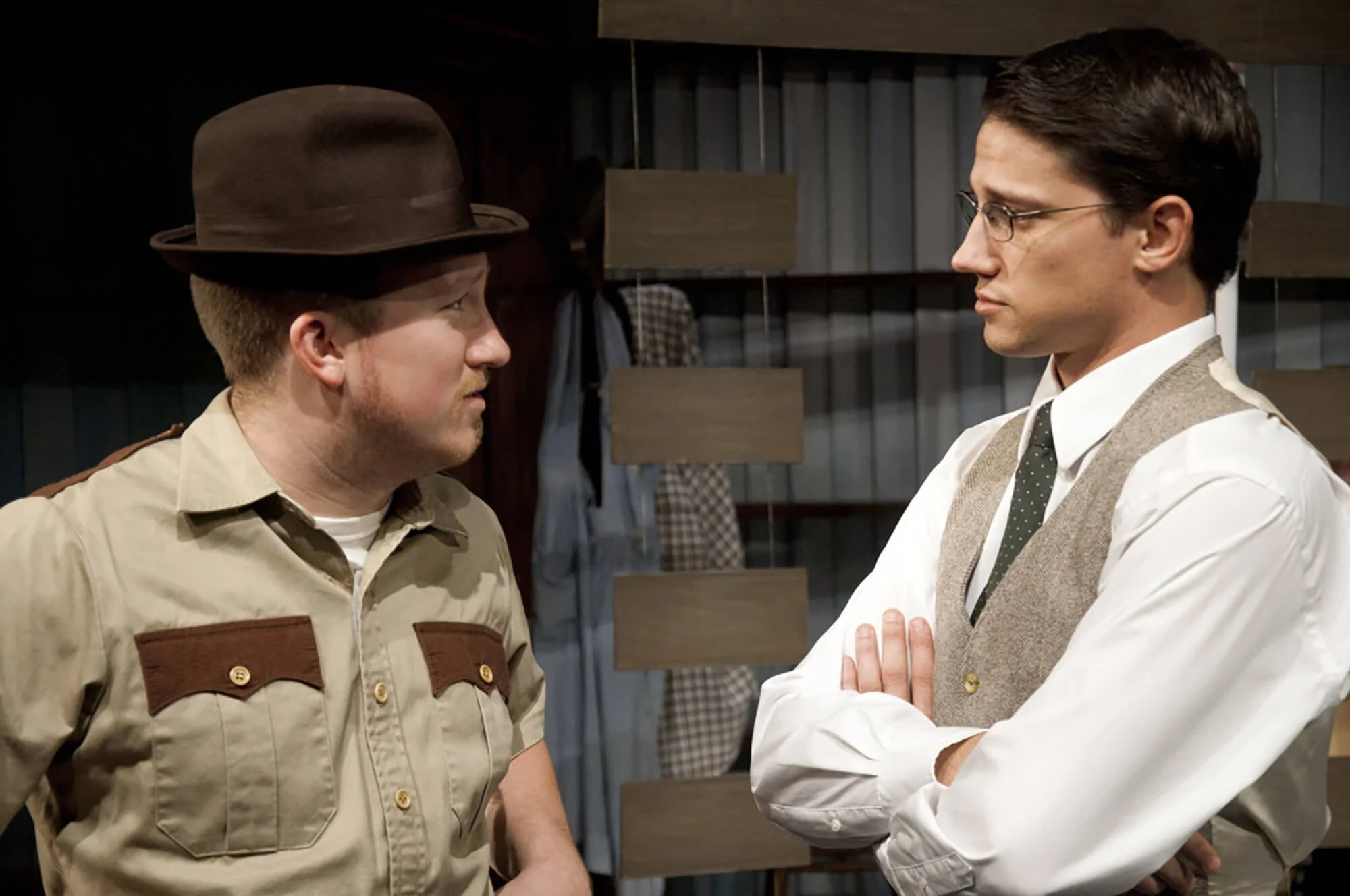 Two men face each other, one dressed as a park ranger and the other as a vintage businessman, in a tense conversation in a wooden cabin or warehouse.