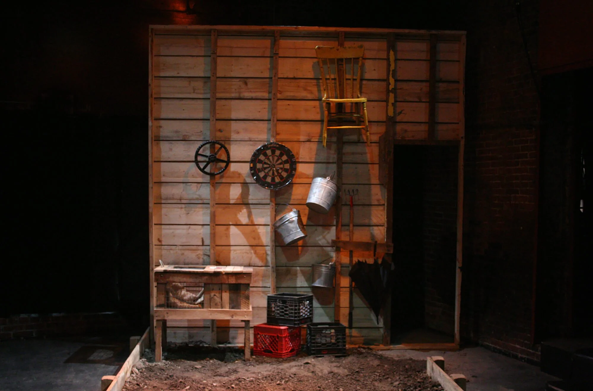 A wooden wall with various objects hanging, including two metal buckets, two crates, a rolling pin, and a wooden chair on top of the wall. There is also a dartboard and a wheel attached to the wall.