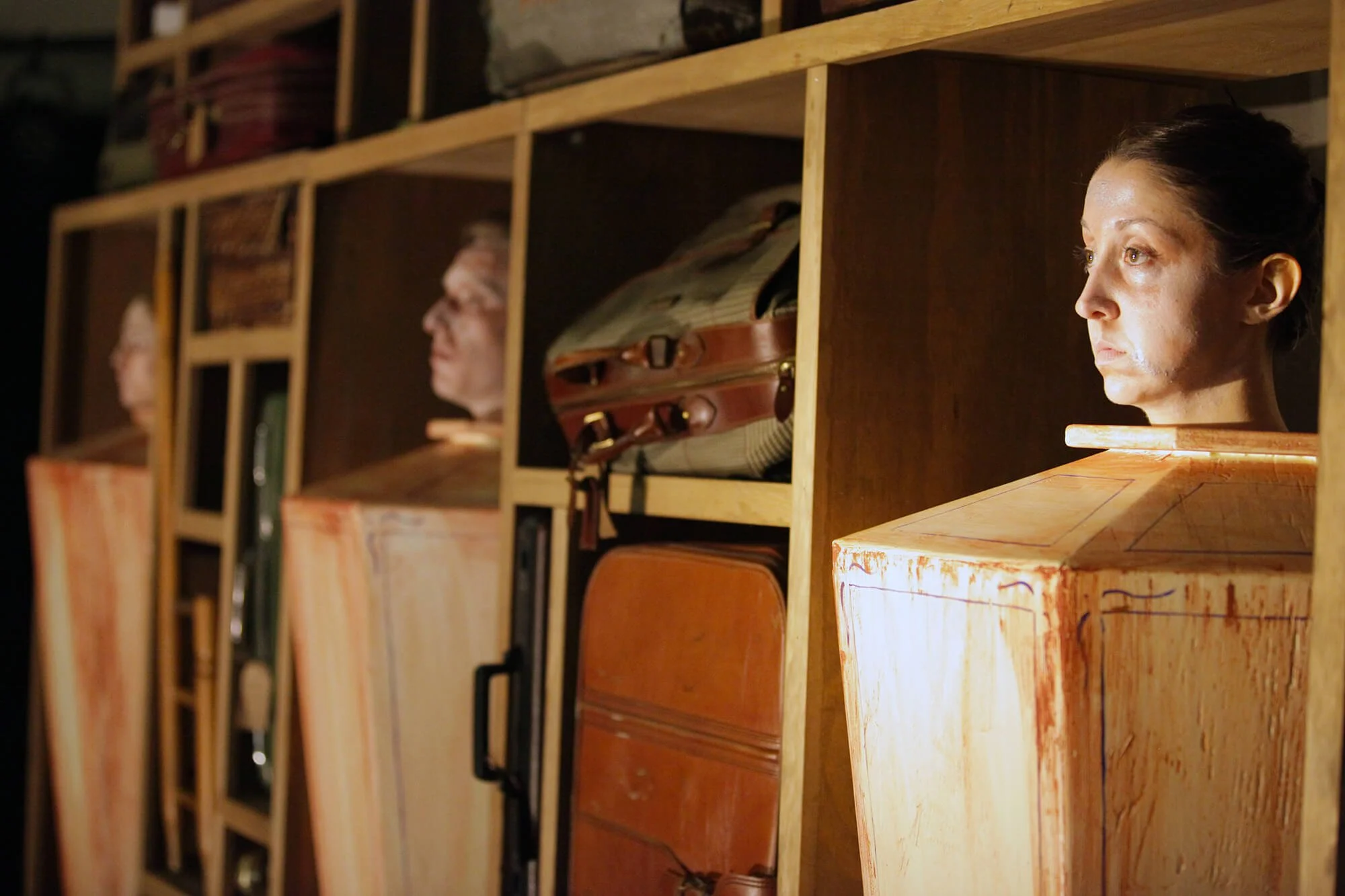 Two women and a man with heads inside wooden cubicles, with a woman looking out from the cubicle on the right.