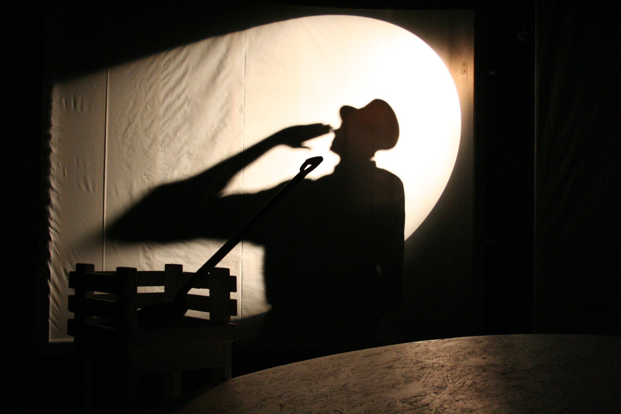 Shadow of a person wearing a hat and holding out a hand, cast on a wall illuminated by a large light source in the shape of a circle, with a piece of furniture in the foreground.