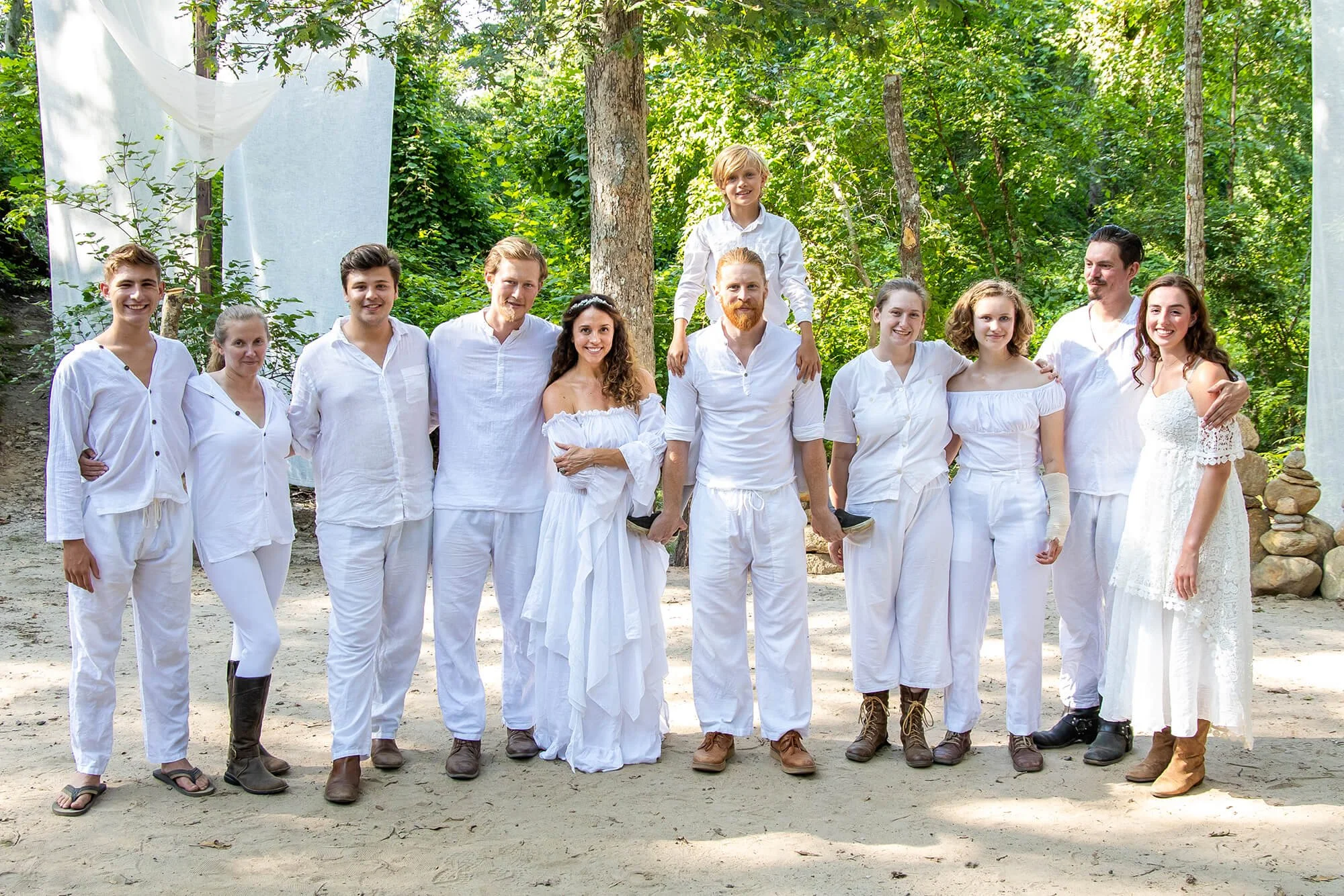 A group of people in white clothing standing outdoors in a wooded area, some with arms around each other, with a young boy sitting on a man's shoulders, smiling.