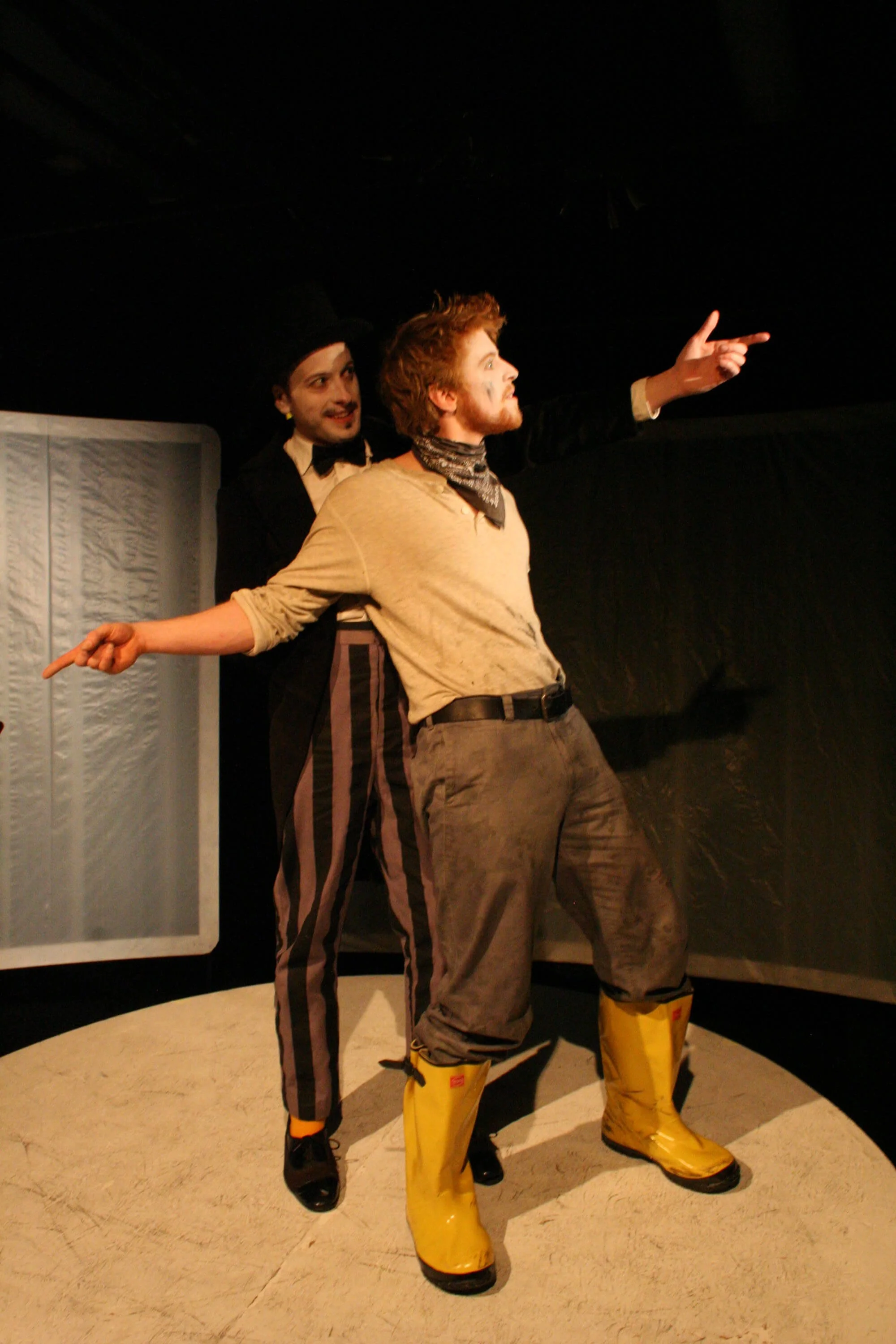Two actors performing on stage, one in black suit and striped pants, the other in a beige shirt and yellow boots, with the man in the beige shirt pointing outward.