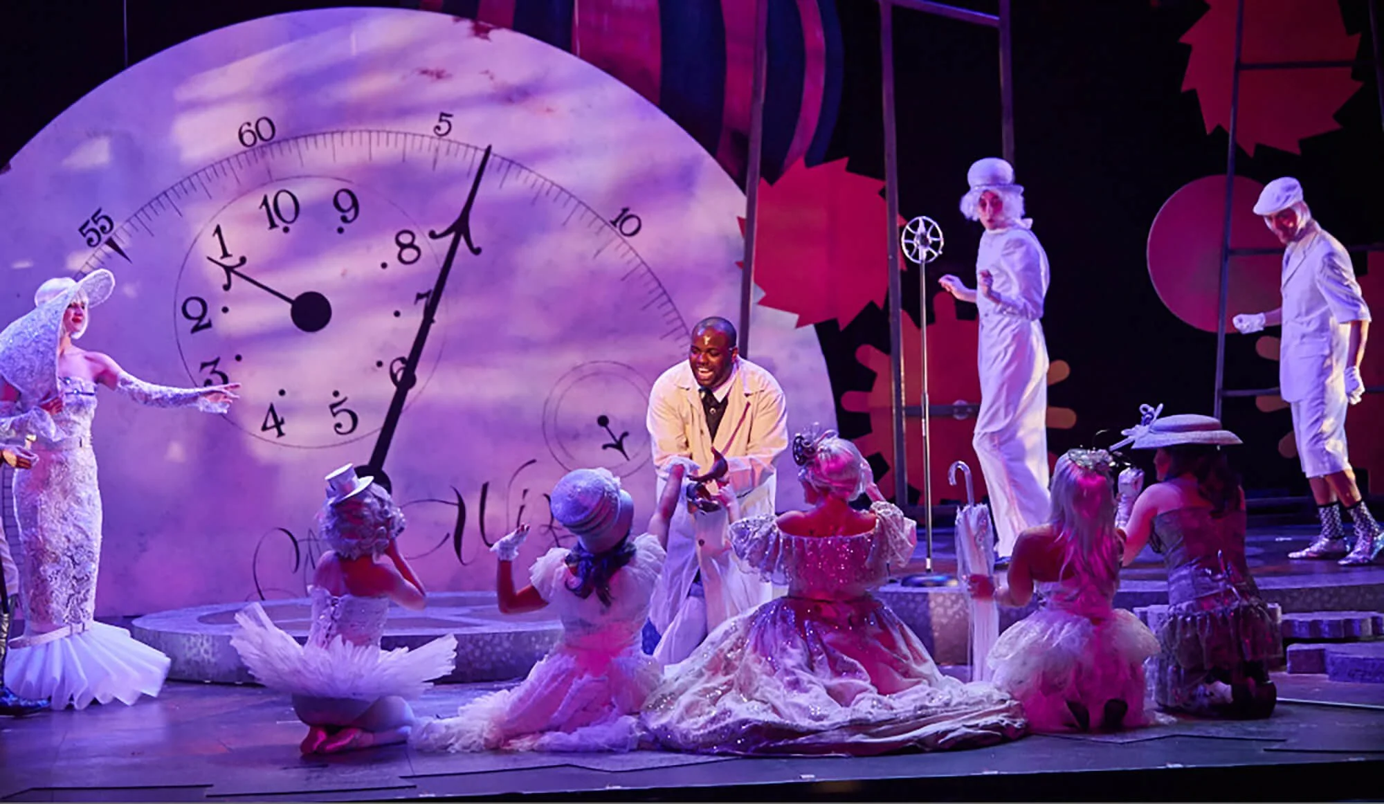 Theatrical stage scene with a large clock in the background and several performers in elaborate costumes, some kneeling or sitting on the floor, others standing, engaged in a lively performance.