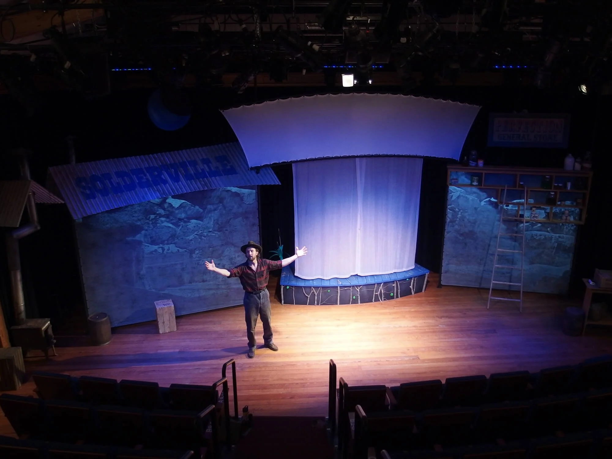 A man is performing on stage with open arms, behind him is a set with trees and mountain scenery and a white curtain, and the stage has wooden flooring and theater seats in the foreground.