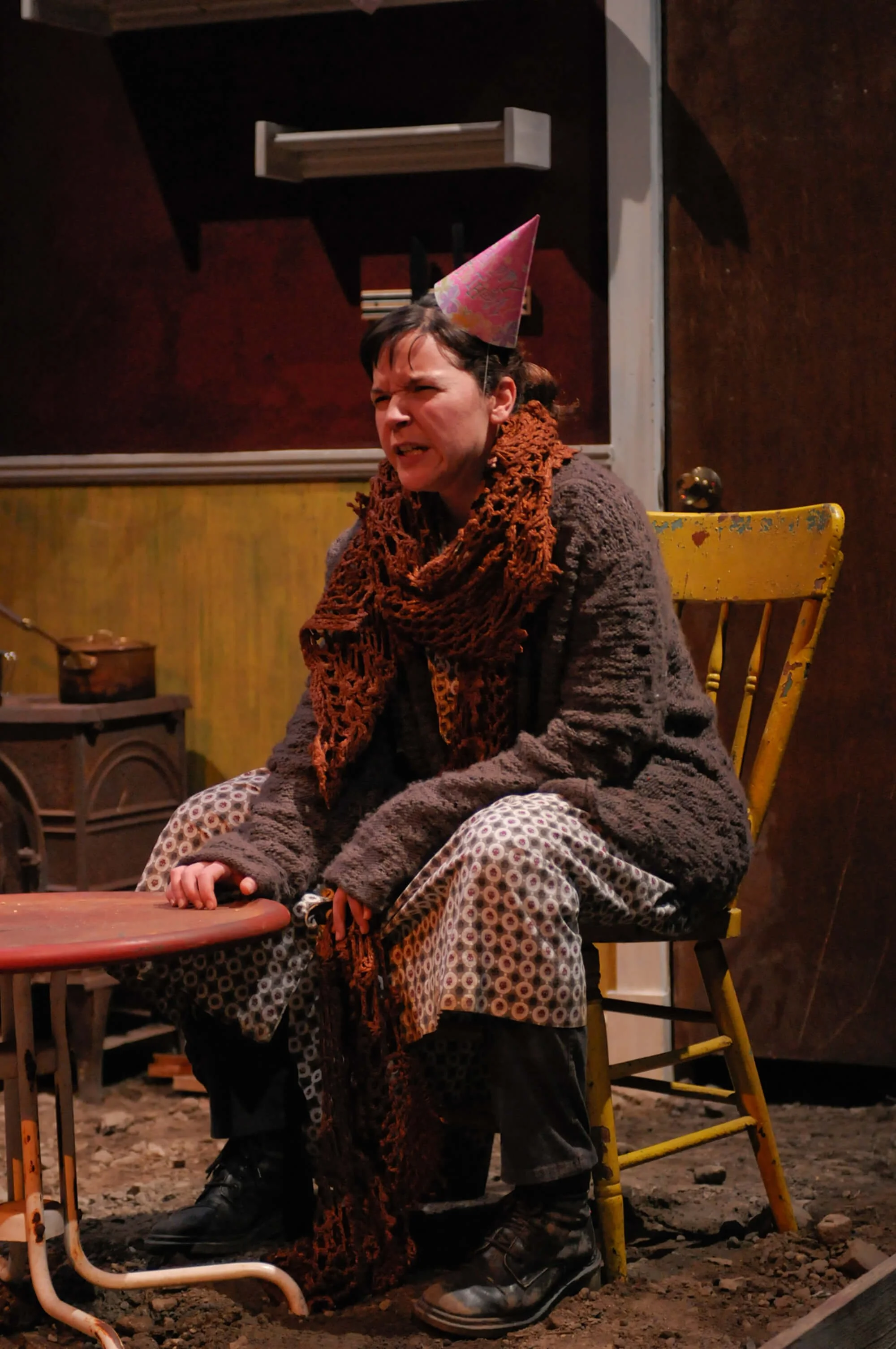 A woman wearing a party hat, scarf, and cozy clothes sitting on a yellow chair, making a grimace or angry face, in a cozy indoor setting with rustic decor.