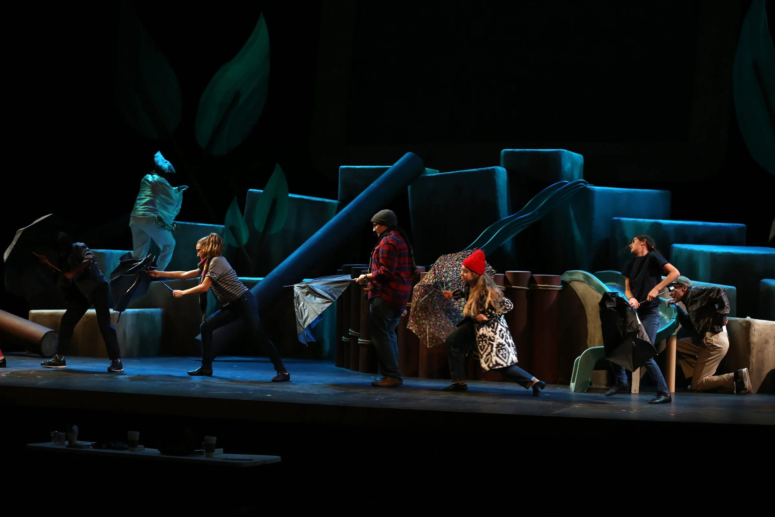 People on stage in a theatrical production, holding umbrellas, with a colorful set background resembling oversized blocks and large leaves.
