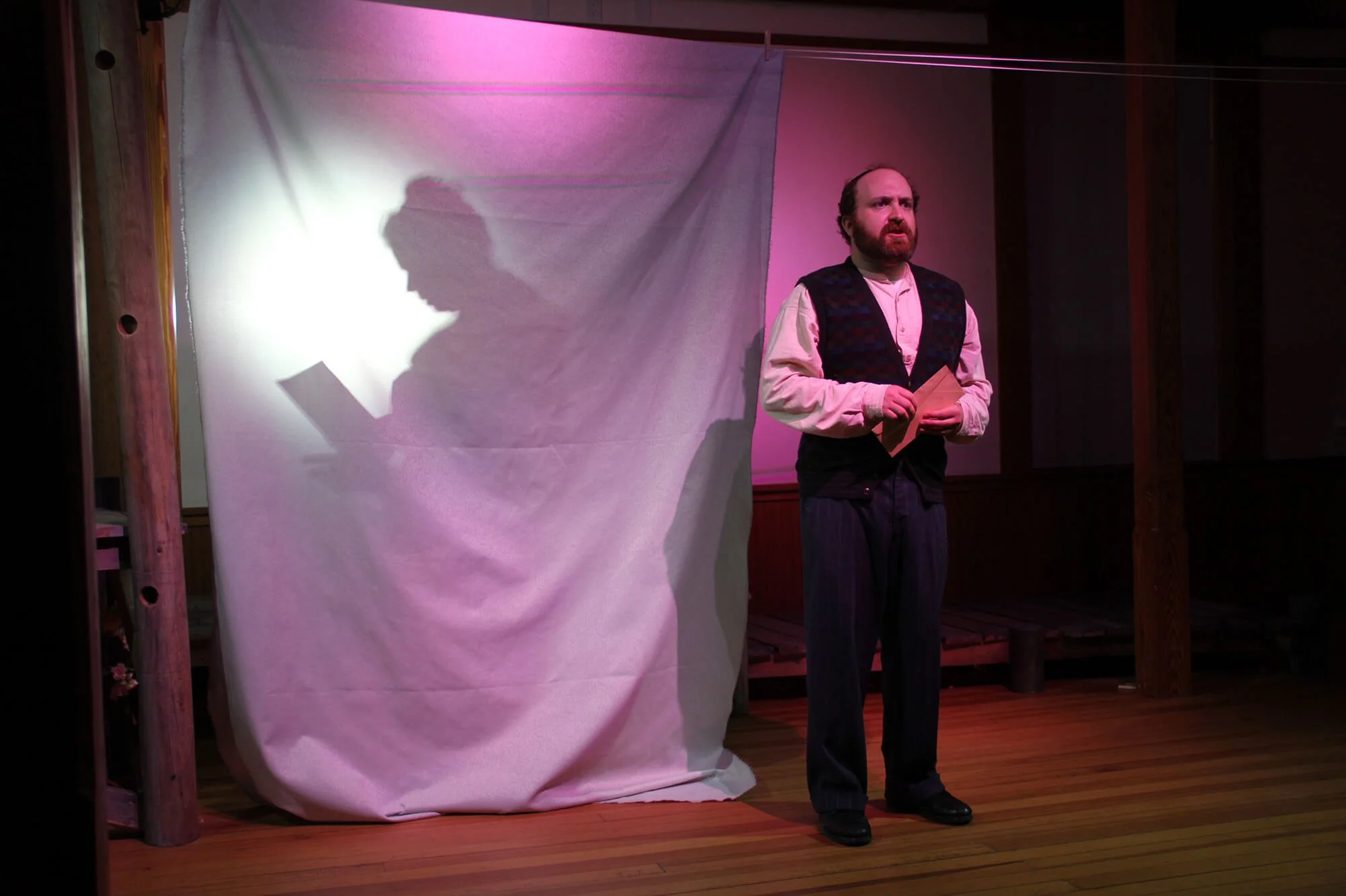 A man with a beard and dark hair standing on a wooden stage, holding a paper, with a white backdrop casting his shadow on it, illuminated by pink stage lights.