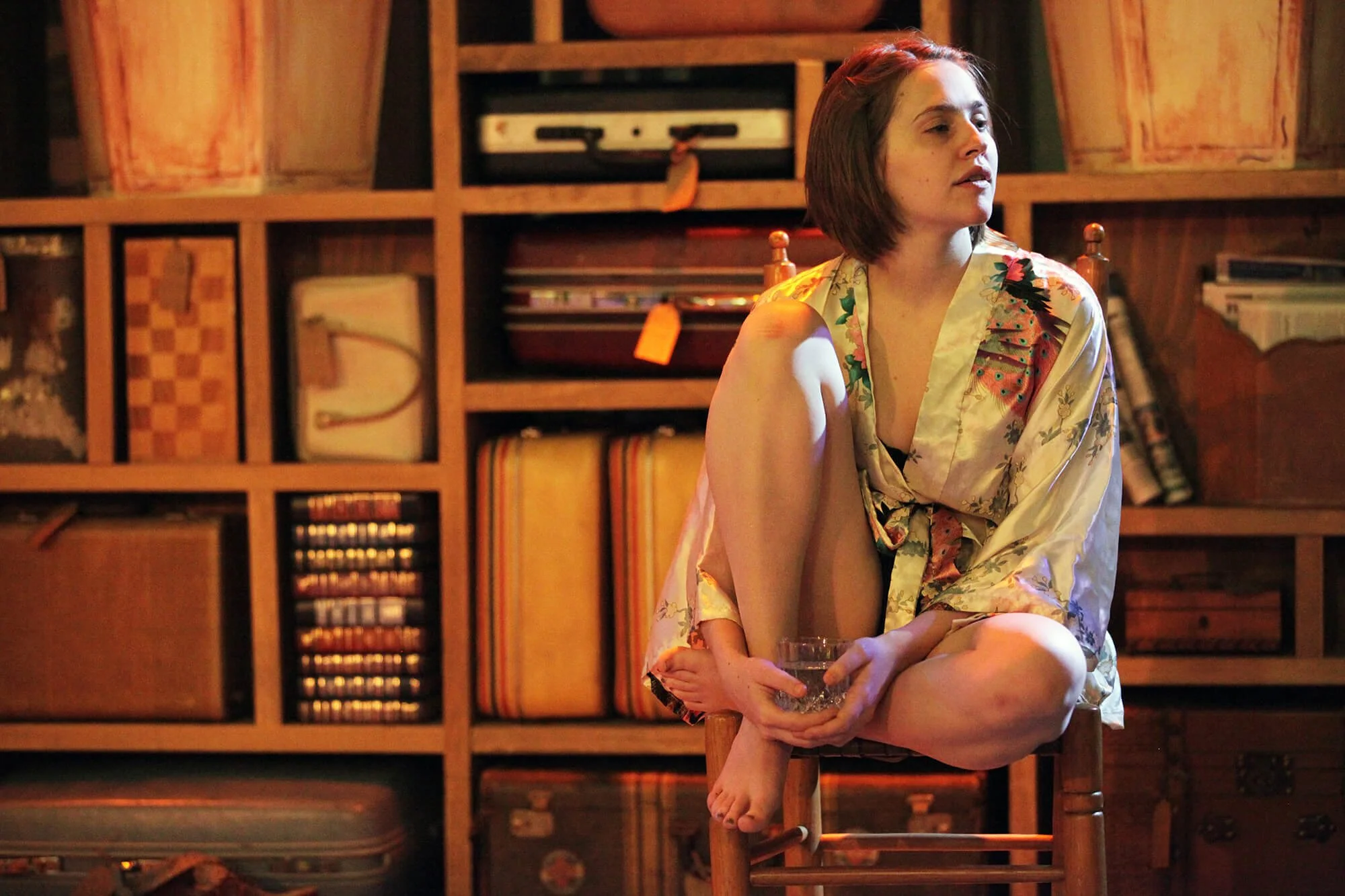 A woman sitting on a wooden chair with one leg bent and holding a glass of water, wearing a silky floral robe in front of a wooden bookshelf filled with books and decorative items.