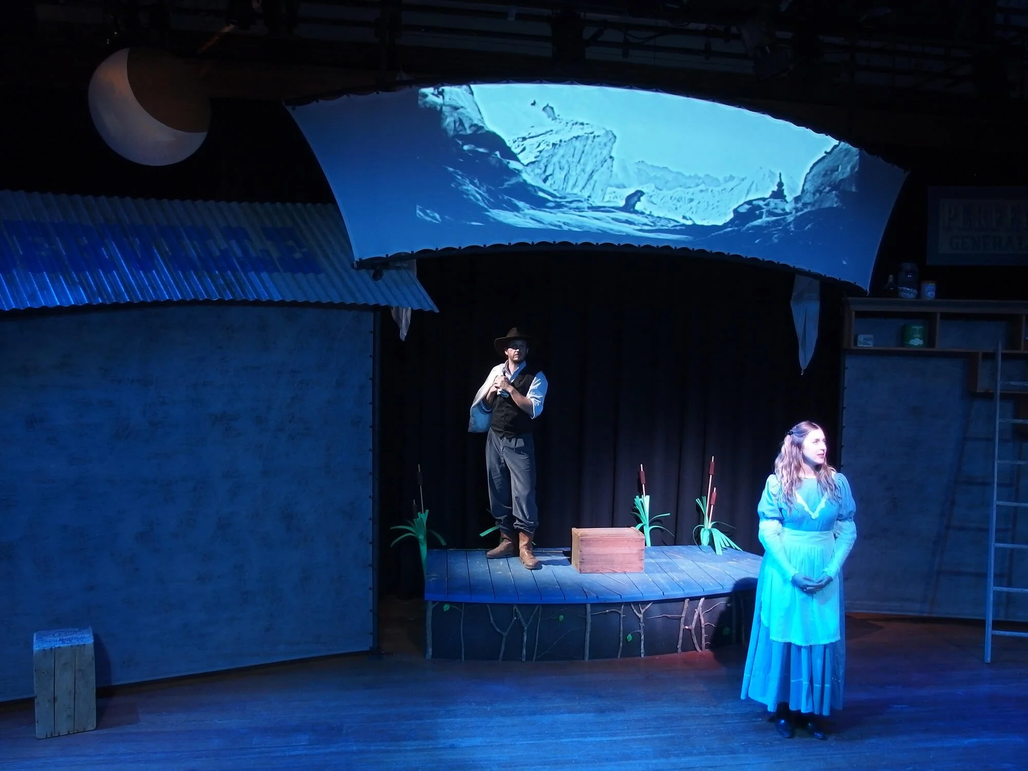 Scene from a theatrical play featuring two actors on stage, one man standing with a mountain backdrop and one woman in a light blue dress standing in front, with stage decorations like plants and a box.