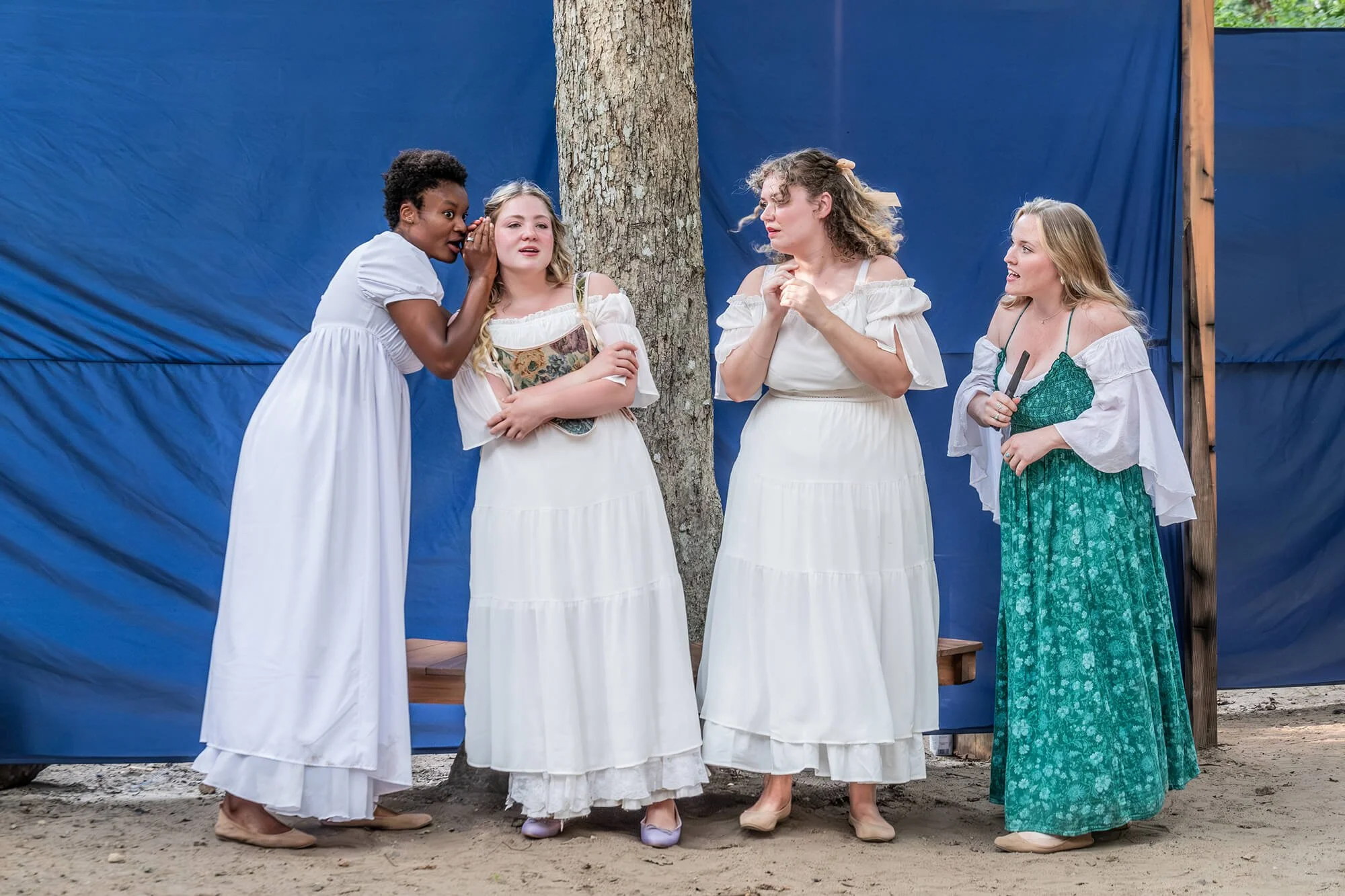 Four women dressed in period costumes gather outdoors around a tree, apparently performing or rehearsing a play.