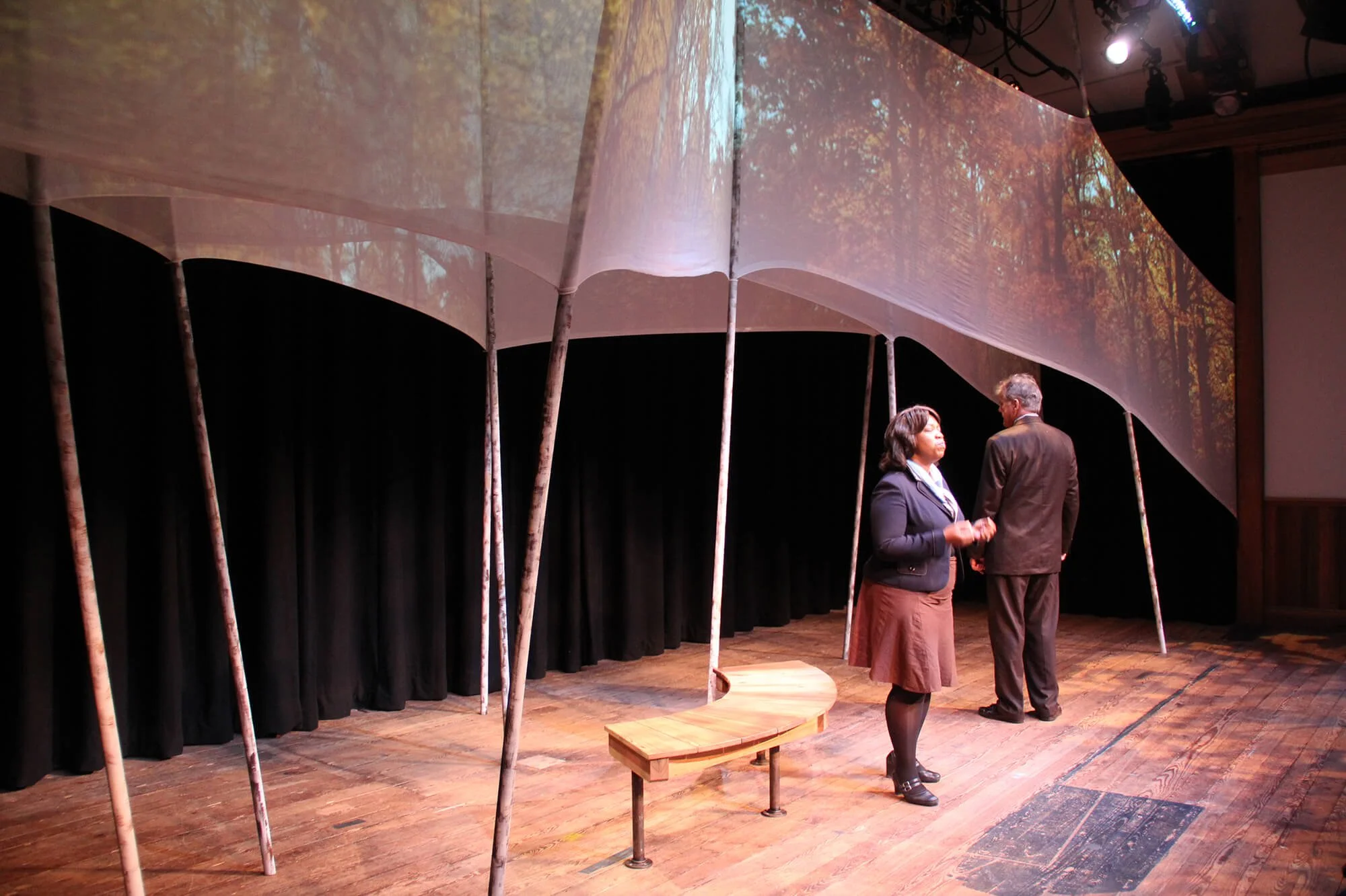 Two individuals are on a stage with a backdrop display of a forest scene. A wood bench sits on the stage, which has a wooden floor. The scene appears to be in a theater or auditorium.