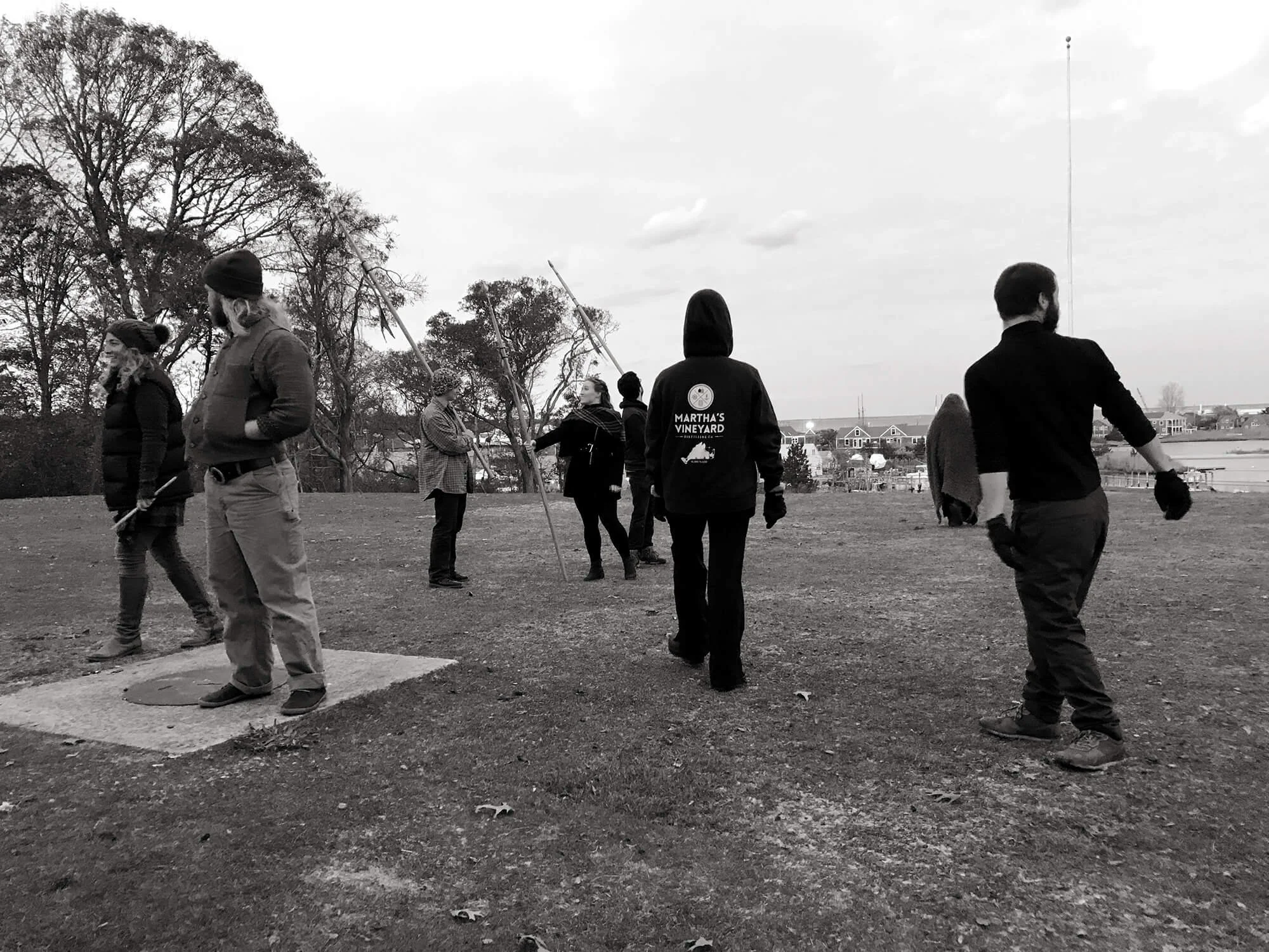 People standing outside on a grassy field, some holding long poles, with trees and houses in the background, in black and white.