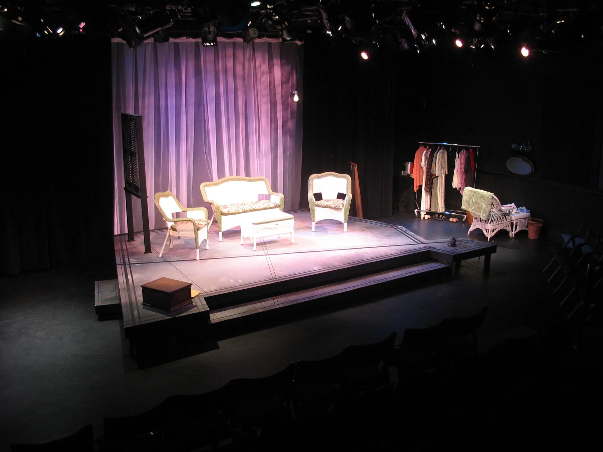 The stage is set with vintage-style white and gold furniture including a sofa, two armchairs, and a bench. A purple curtain backdrop and a clothing rack with various costumes are on the stage, indicating a theatrical play or performance setting.