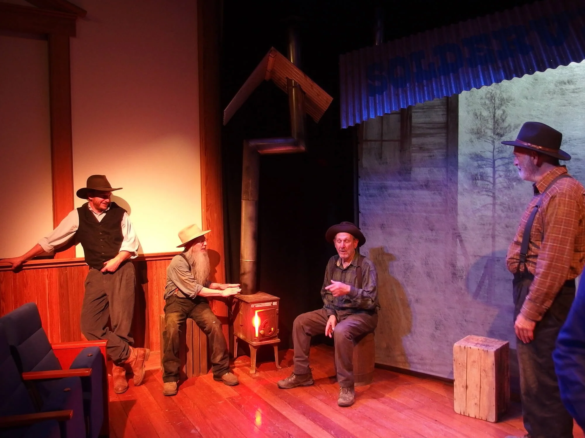 Four men in vintage western costumes on a stage, with one man sitting and the others standing, including a man with a long white beard and a man with a gray hat, in a setting resembling an old western cabin.