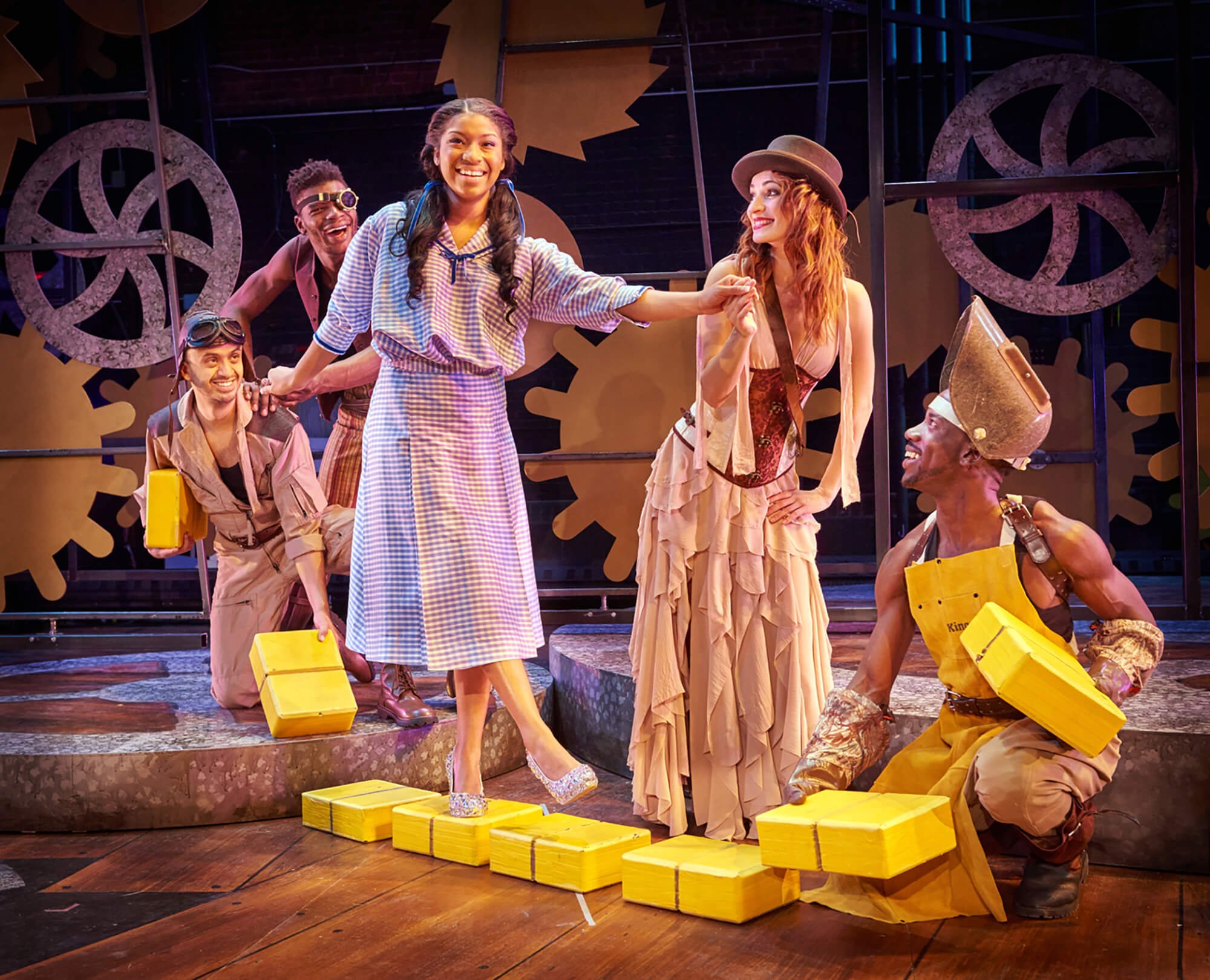 Actors on stage performing a theatrical production with steampunk and adventurer costumes, interacting with yellow props resembling boxes or tools, against a backdrop of gears and cogs.