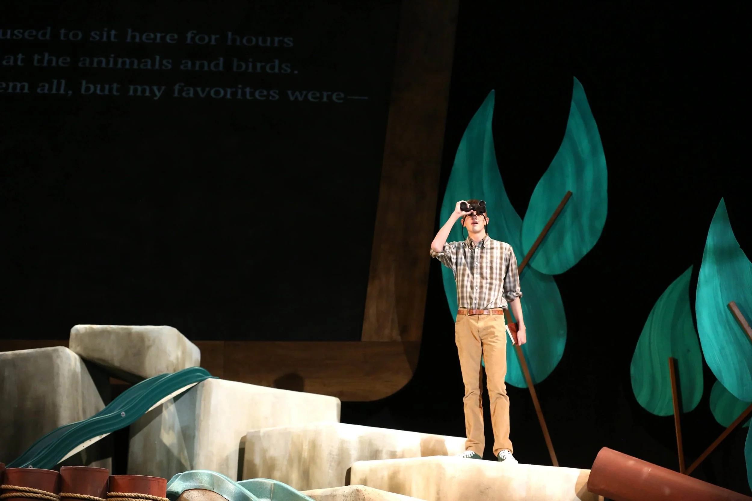 A young man in plaid shirt and khaki pants on a stage using binoculars, with large green foliage decorations in the background and a dark screen with partially visible text.