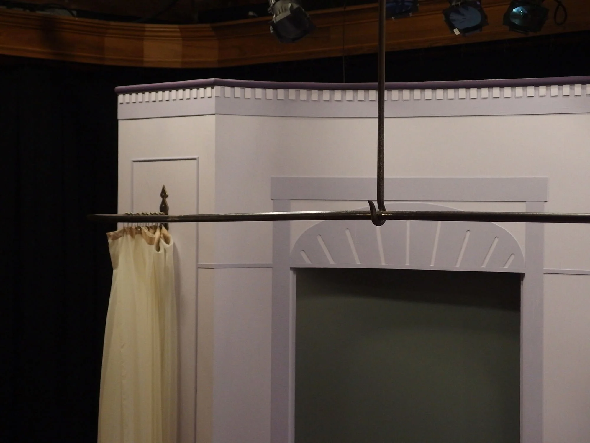 Stage set with a white fireplace, a cream-colored curtain hanging on a black wall, and a metal rod suspended from the ceiling for a theatrical performance.