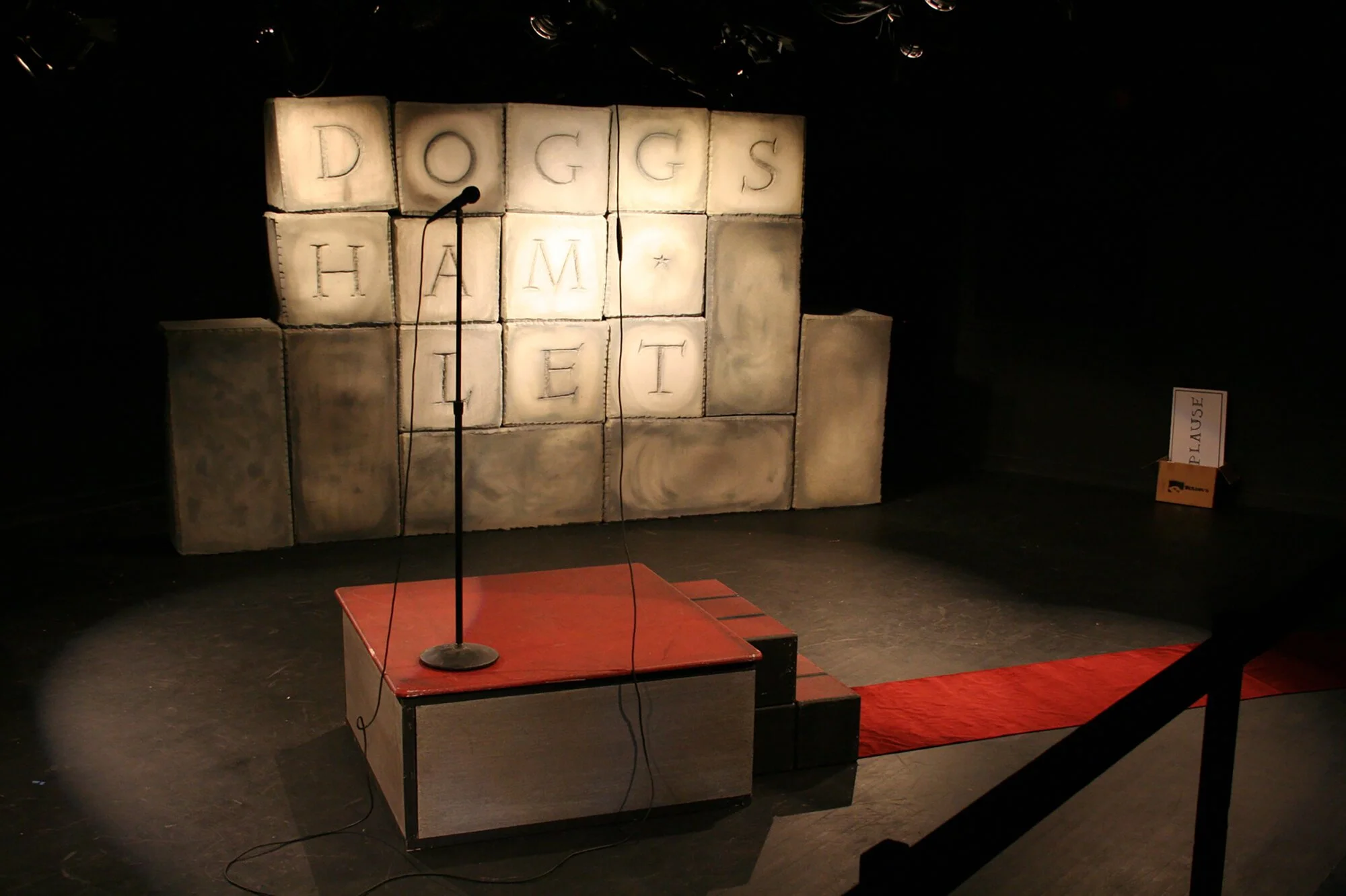 Stage setup with a backdrop made of large cubes with letters spelling 'DOGG'S HAMLET'. There is a red carpet leading to steps and a microphone on a stand in front of the backdrop.