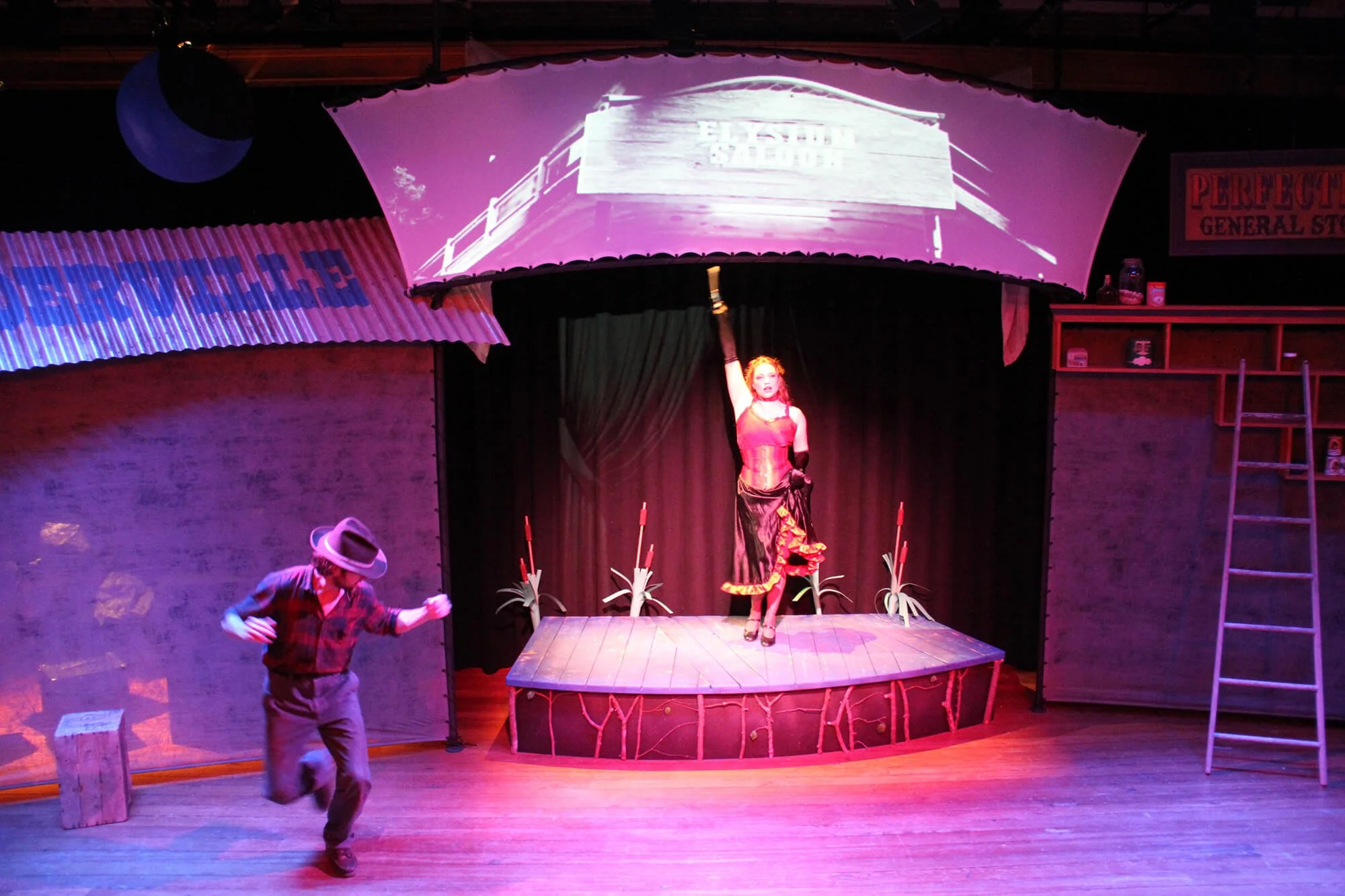 A stage performance featuring a woman in a red and black costume with a long skirt, posing with one arm raised on a small raised platform. There is a child in a hat and plaid shirt dancing to the left in front of the stage. The background has a black