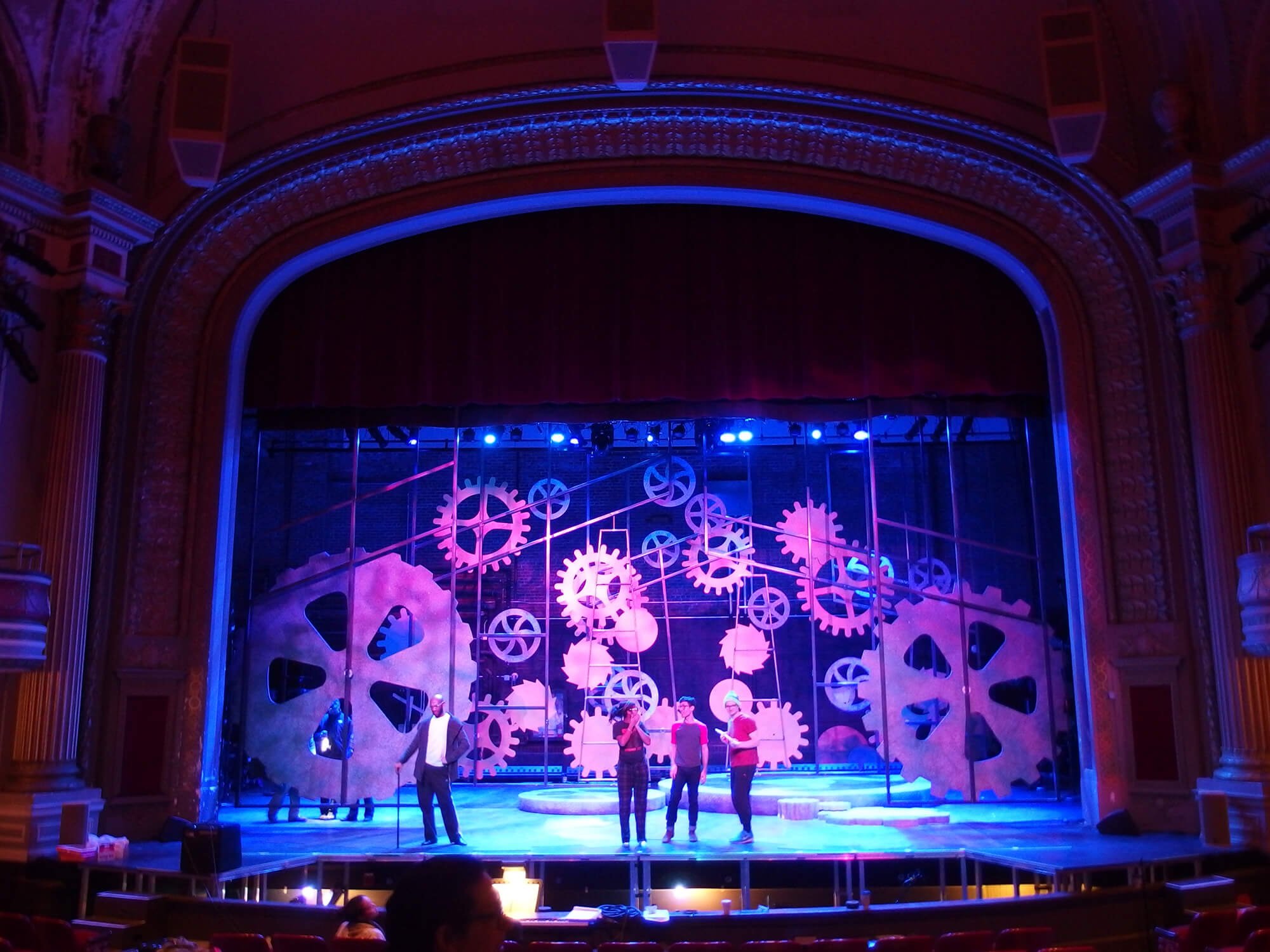 Actors on stage during a theatrical performance with large gear and cogs set design, illuminated with pink and blue stage lighting, in a grand theater with ornate architecture.